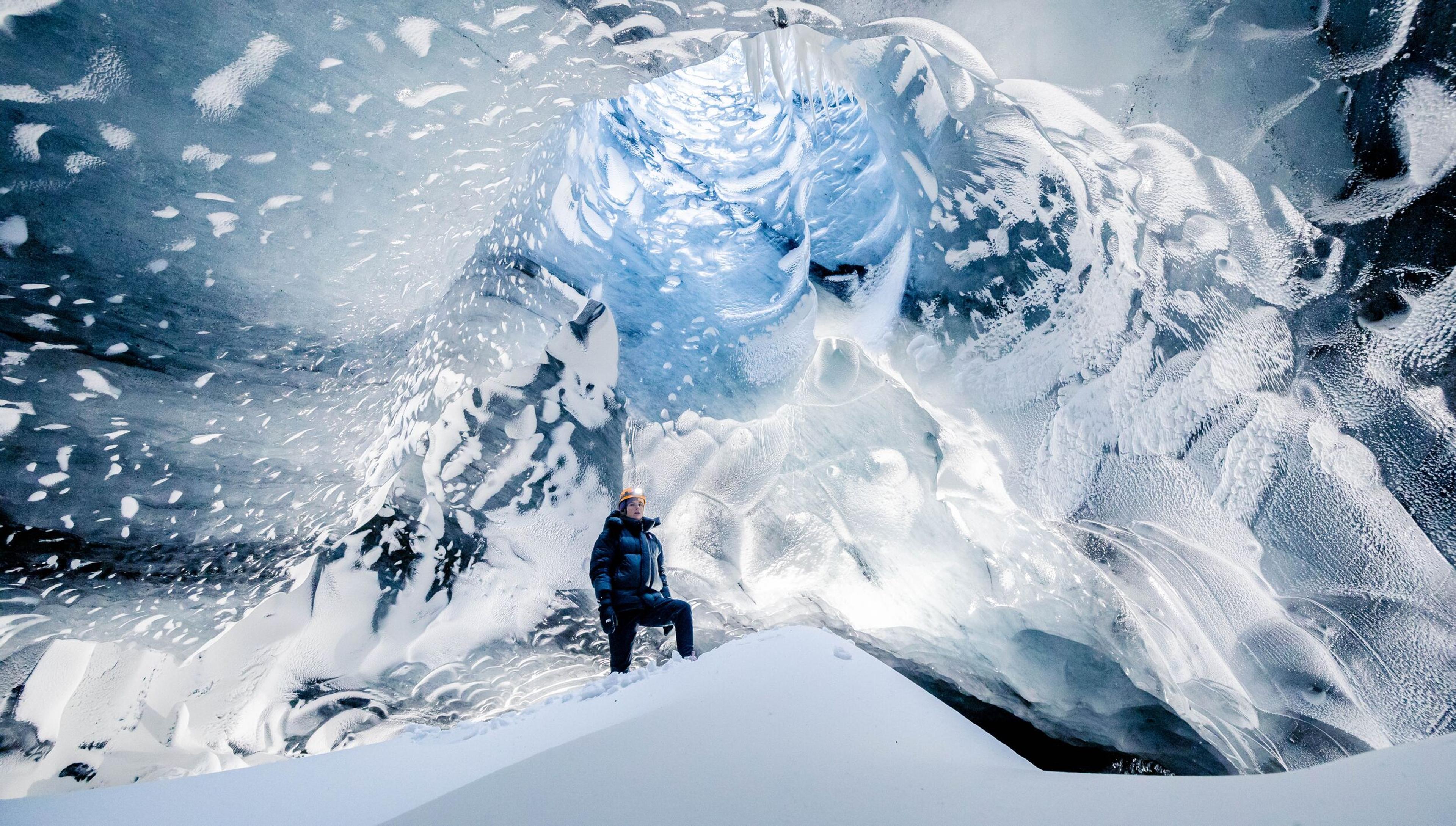 A person standing in front of a large, intricate ice cave with a backdrop of a snow-covered mountain. The cave's walls are textured with layers of snow and ice, resembling a wave above the person, who appears minuscule in comparison.