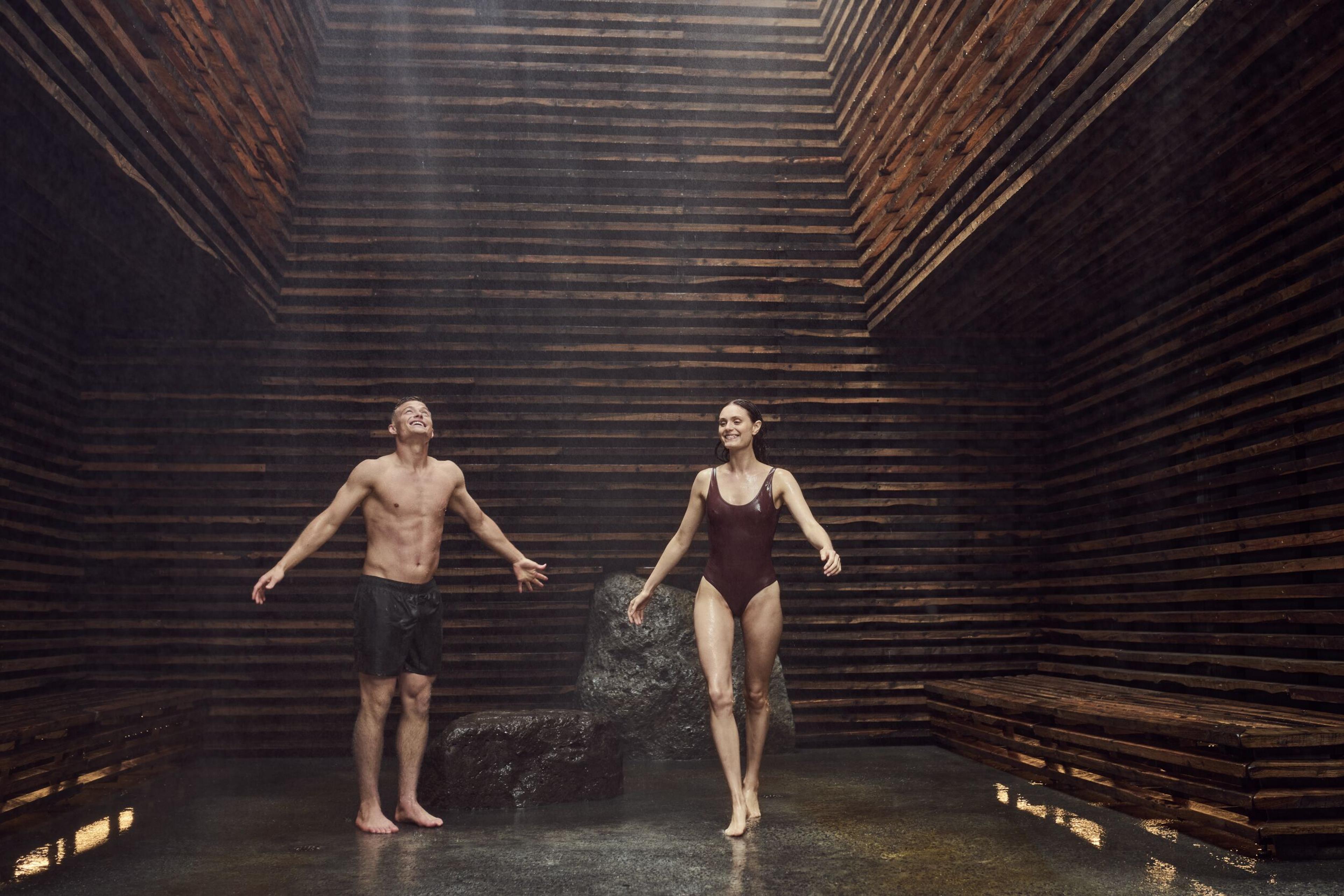 Couple enjoying the cold mist ritual at Sky Lagoon in a wooden, open-roof room with water falling gently from above.