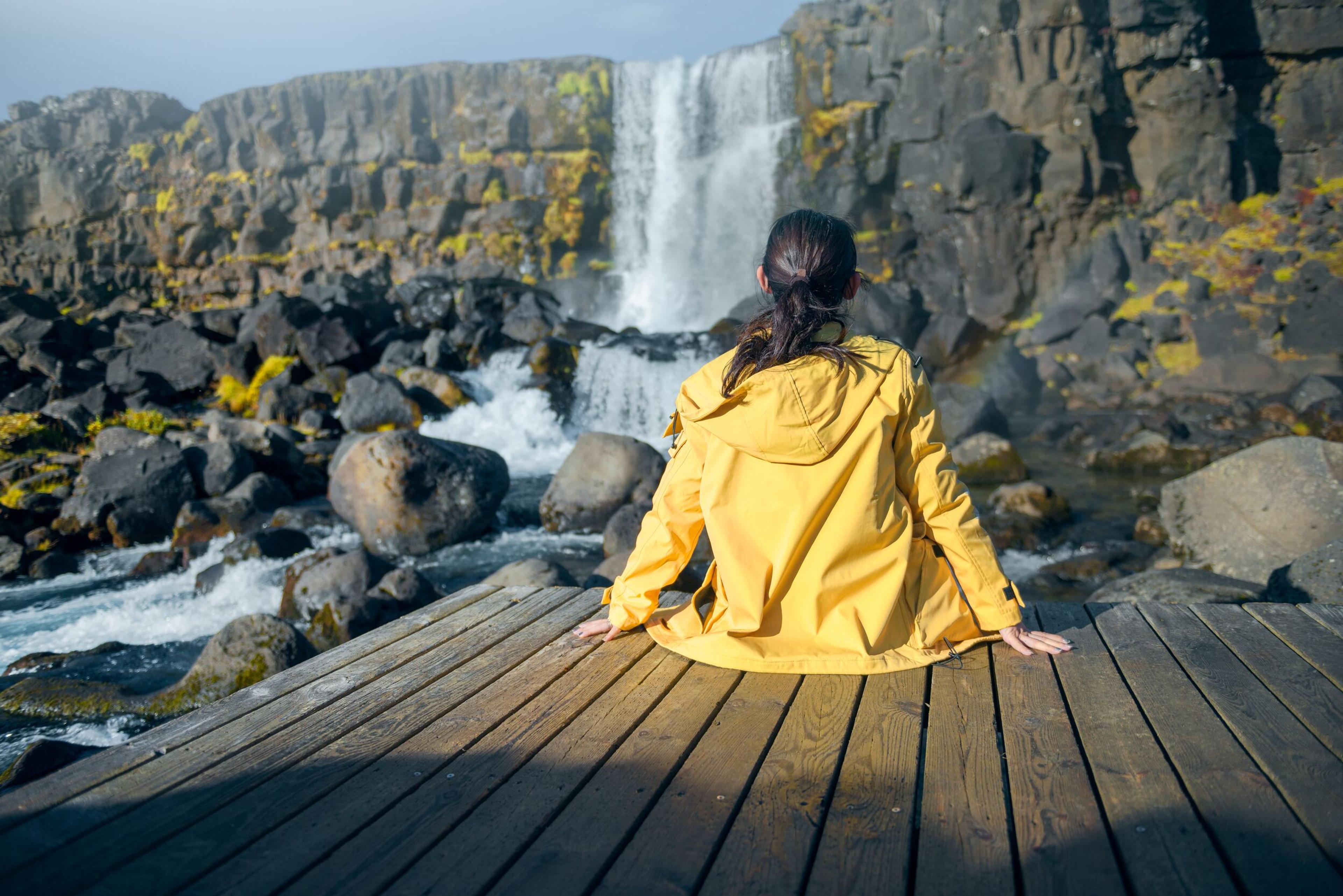 A person in a yellow jacket sitting on a wooden deck, gazing at a waterfall cascading down a rugged cliff amidst a rocky landscape, a moment of peaceful reflection in nature.