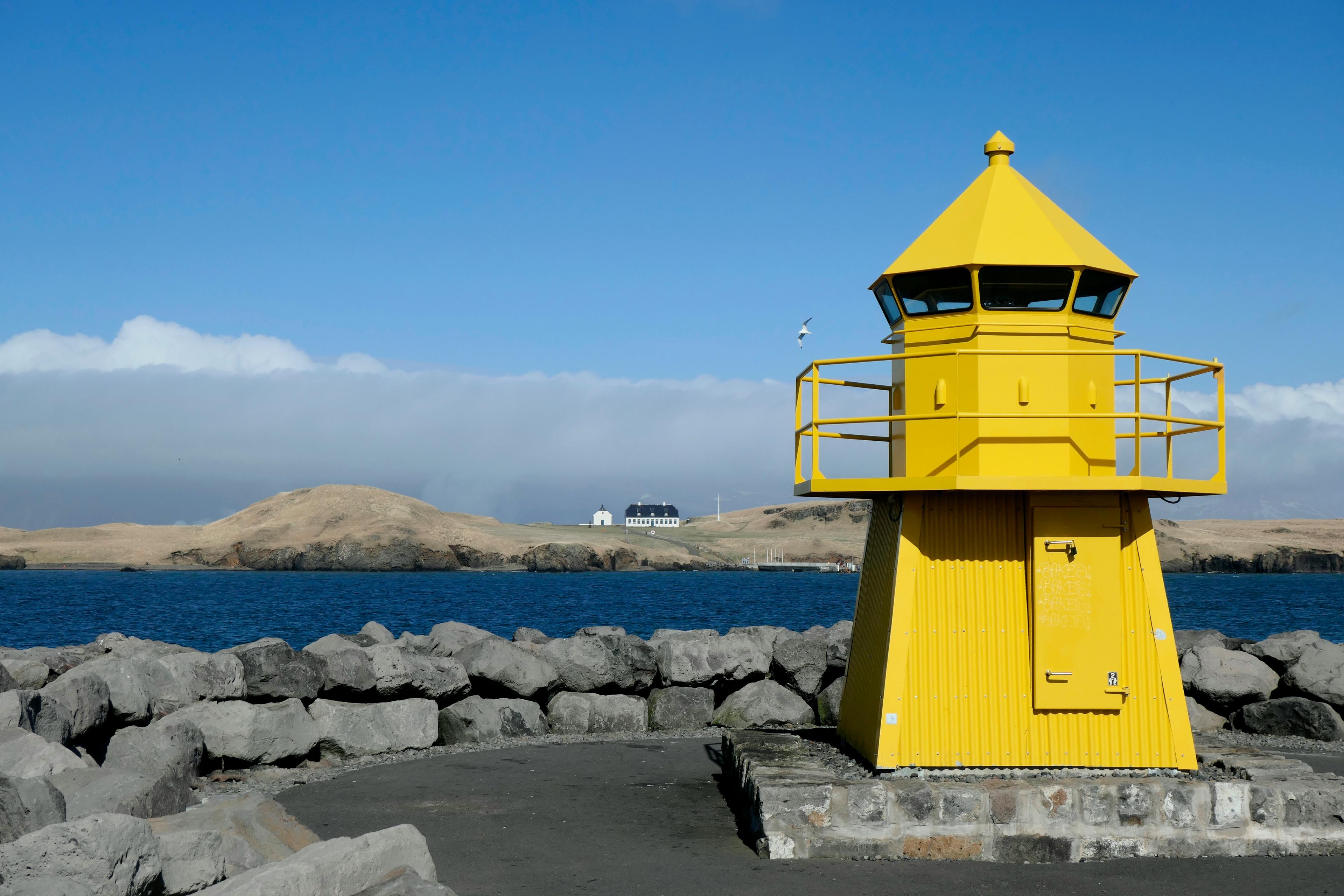 Bright yellow lighthouse on a rocky pier overlooking the ocean and distant hills under a clear blue sky.