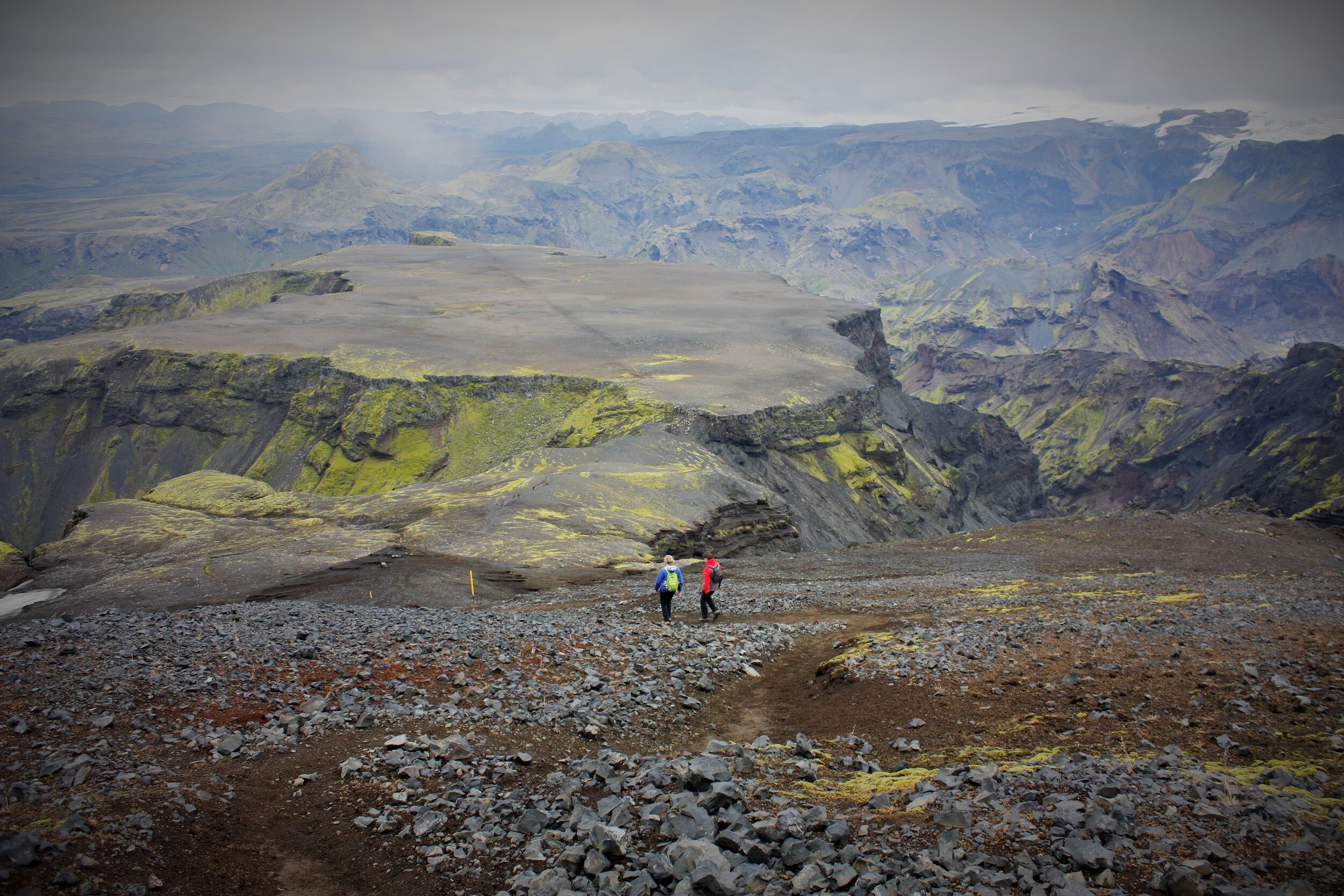 Two hikers walk along a rocky path through volcanic terrain with moss-covered cliffs in the Iceland Highlands.