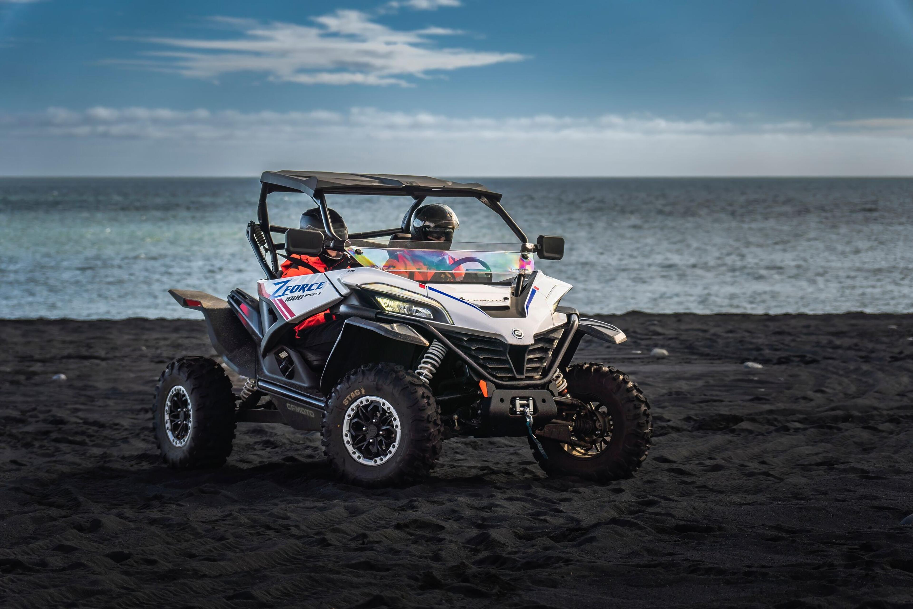 Two people in orange gear with a black off-road ATV on a rugged black sand terrain in Iceland.