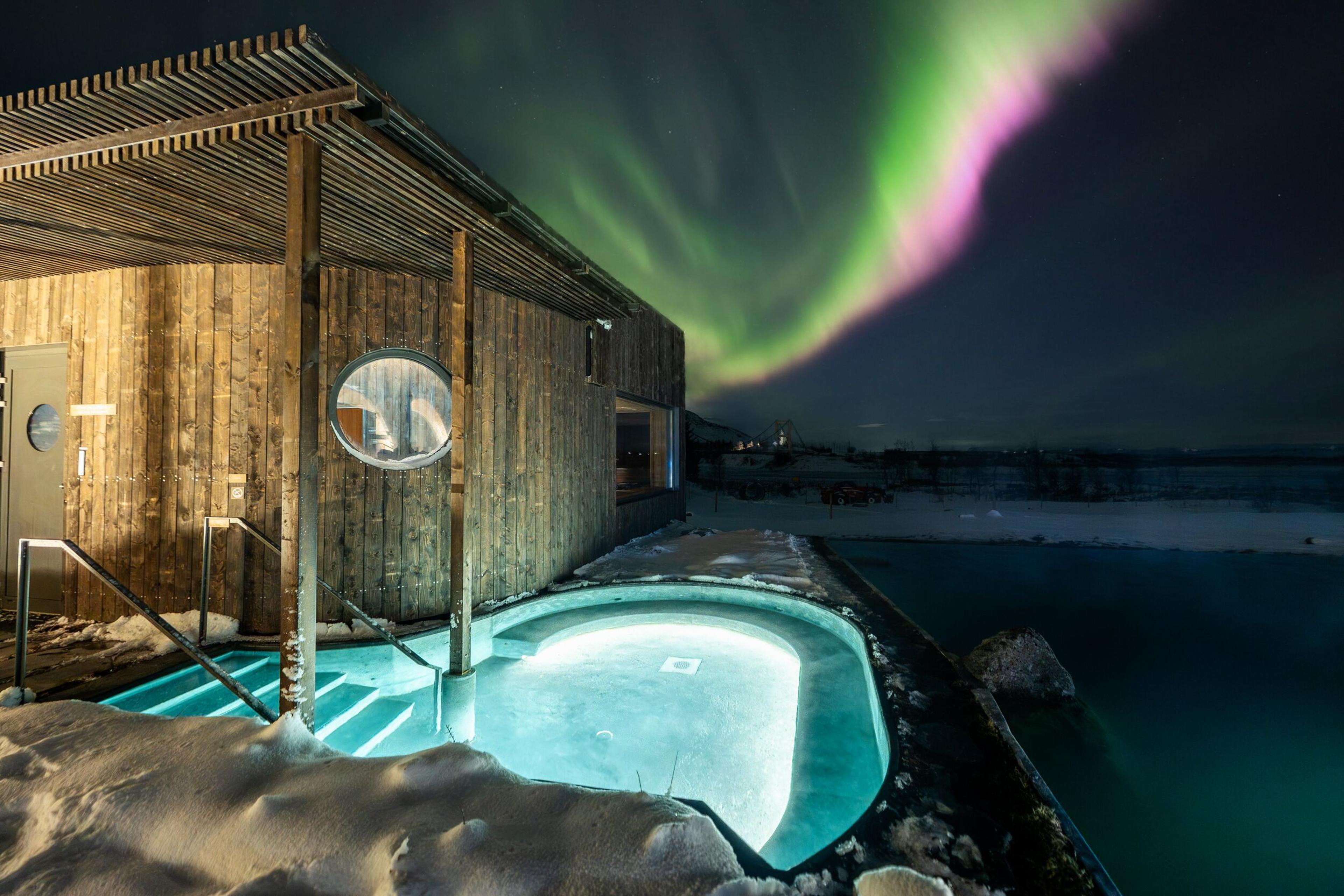 Outdoor spa with a hot tub and wooden building under vibrant green and pink Northern Lights in a snowy landscape.