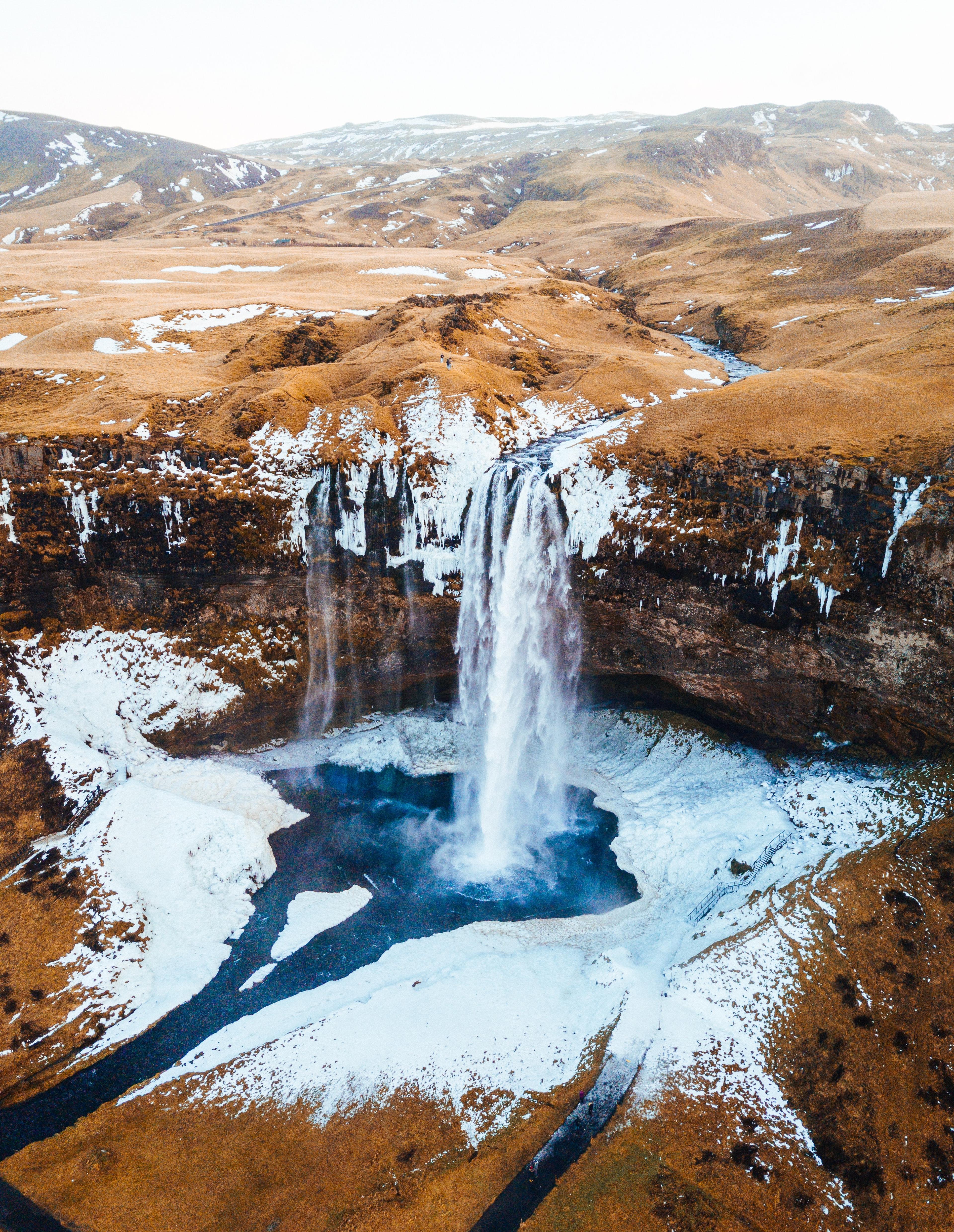 Aerial perspective of Seljalandsfoss waterfall with scattered frozen patches amidst a brown, snow-free landscape.