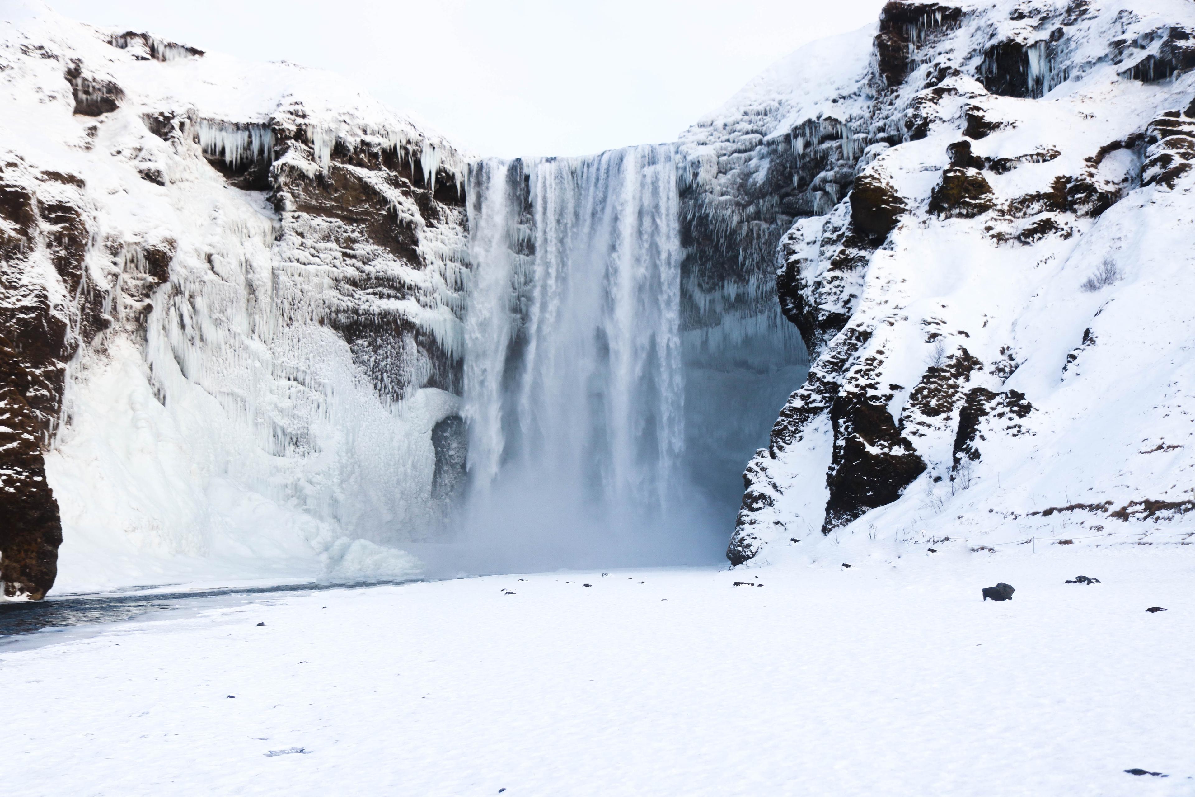 a majestic waterfall in winter, with water cascading down from a height and a cloud of mist rising from where the water meets the frozen river below. The waterfall is flanked by ice-covered cliffs, and the surrounding landscape is blanketed with snow