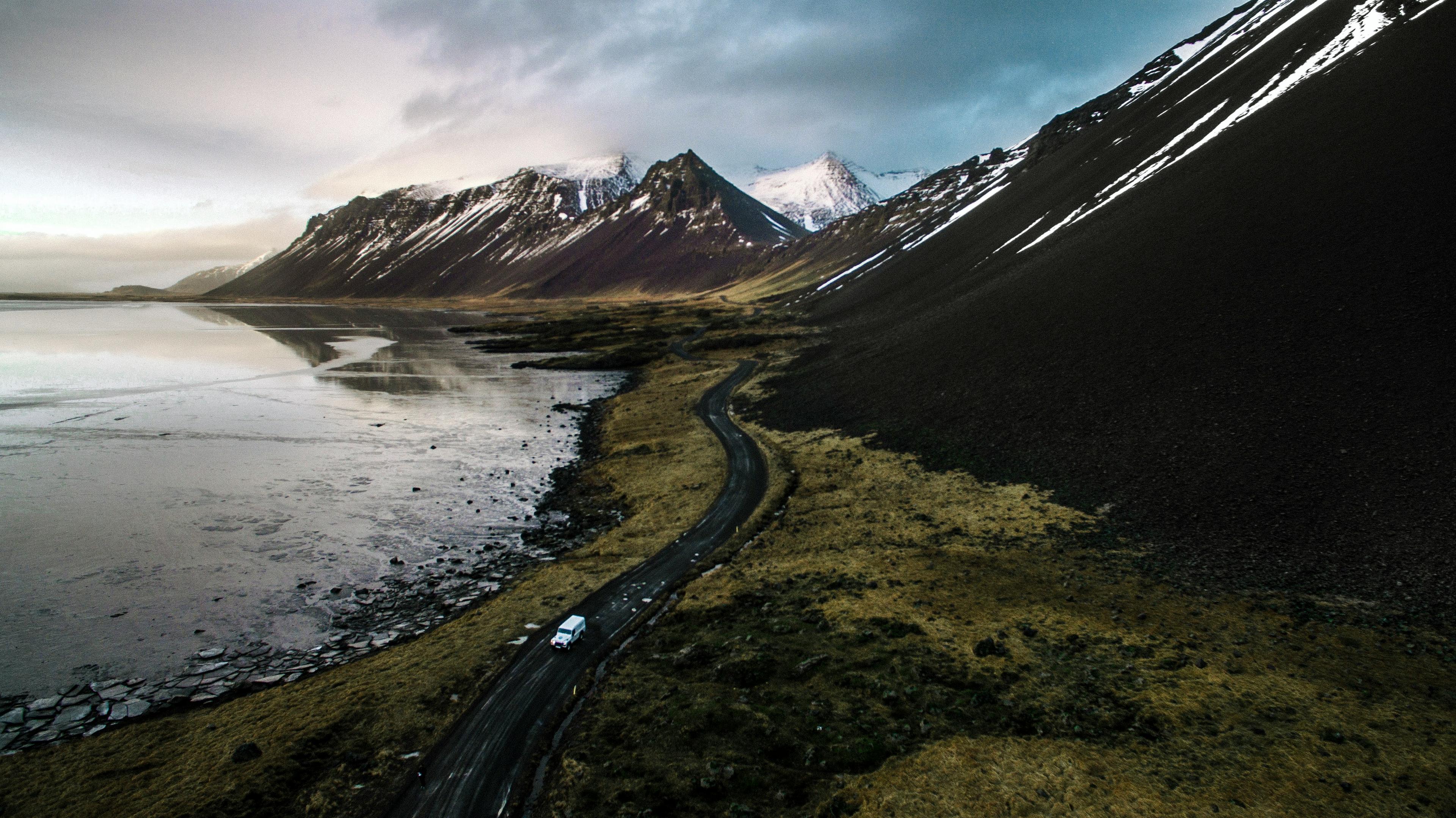  Aerial view of a car driving on a winding coastal road near snow-capped mountains in Iceland, showcasing scenic landscapes.