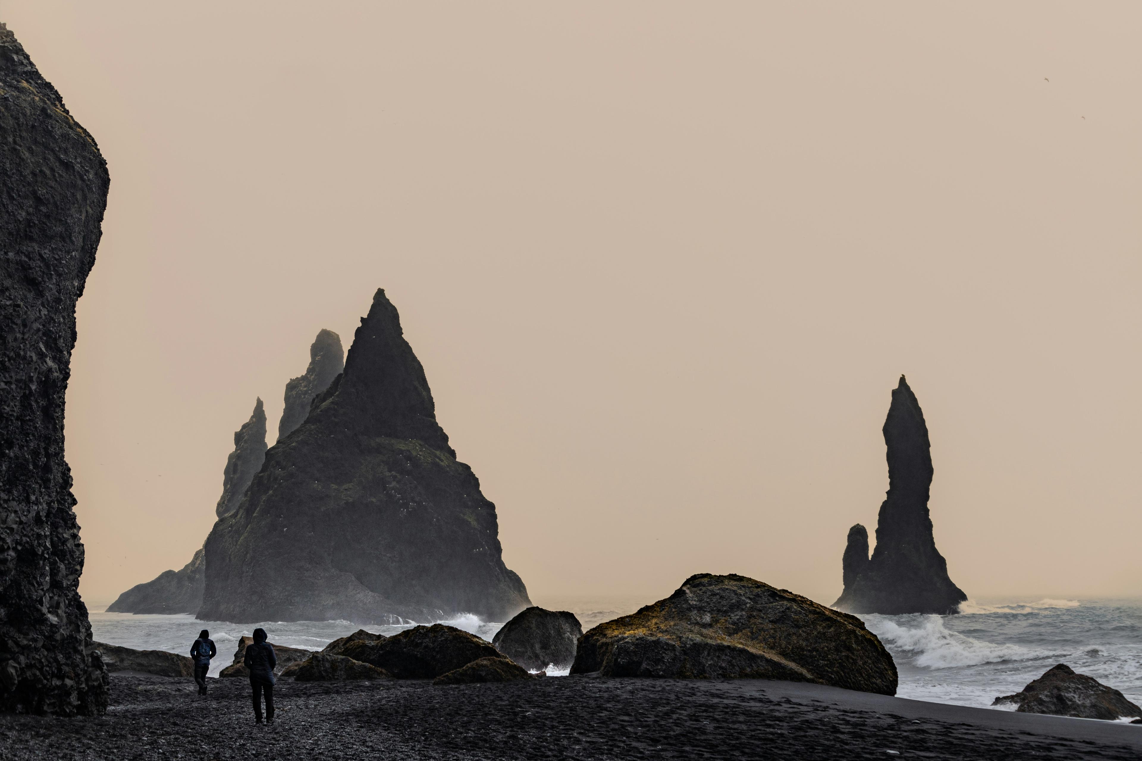 This image showcases Reynisfjara Beach in Iceland, featuring its iconic basalt sea stacks rising sharply from the water. The black sand beach contrasts with the pale, overcast sky, and two small figures can be seen walking near the towering formations, emphasizing their immense scale. 