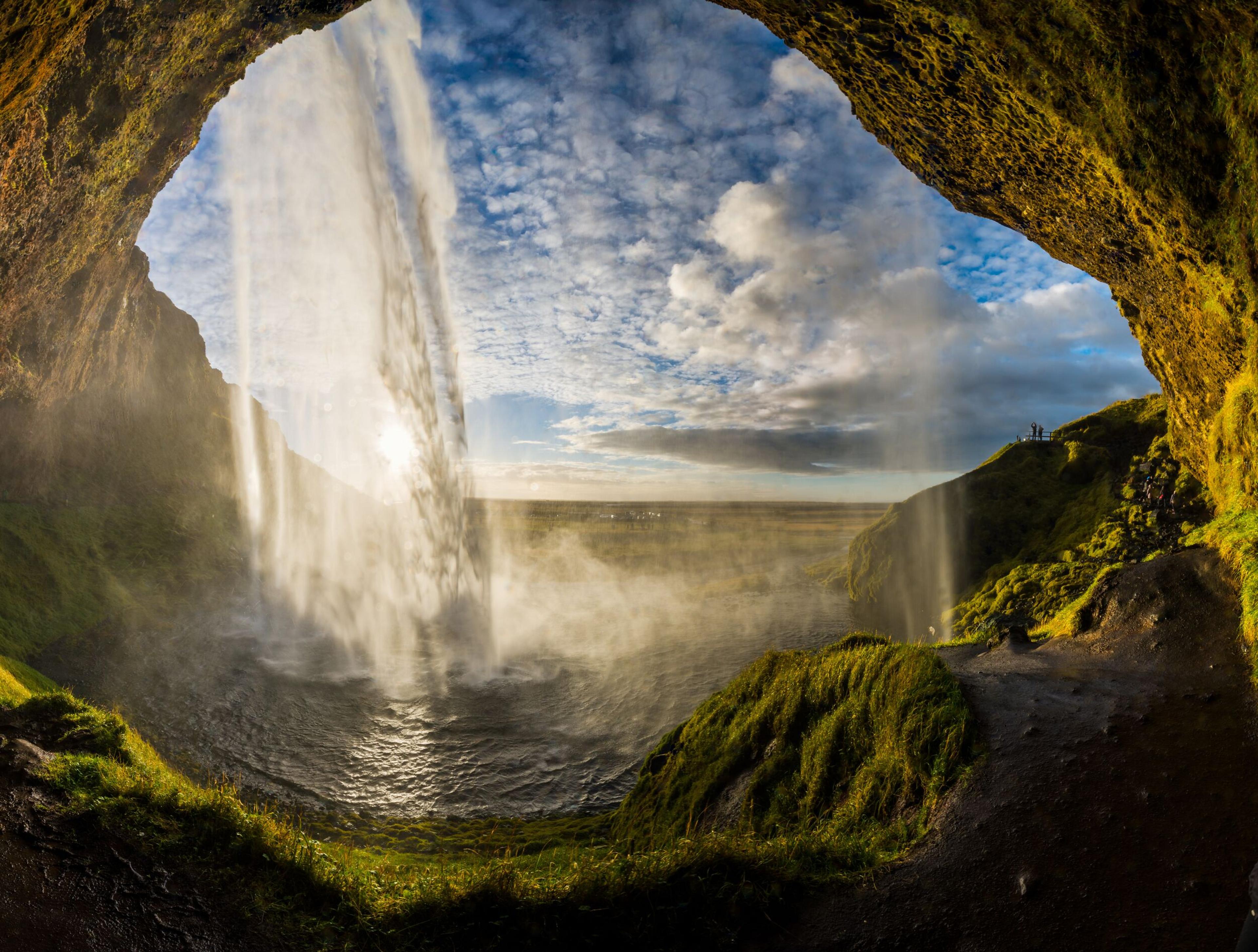 Waterfall viewed from behind inside a mossy cave, with a misty green landscape under a cloudy sky.