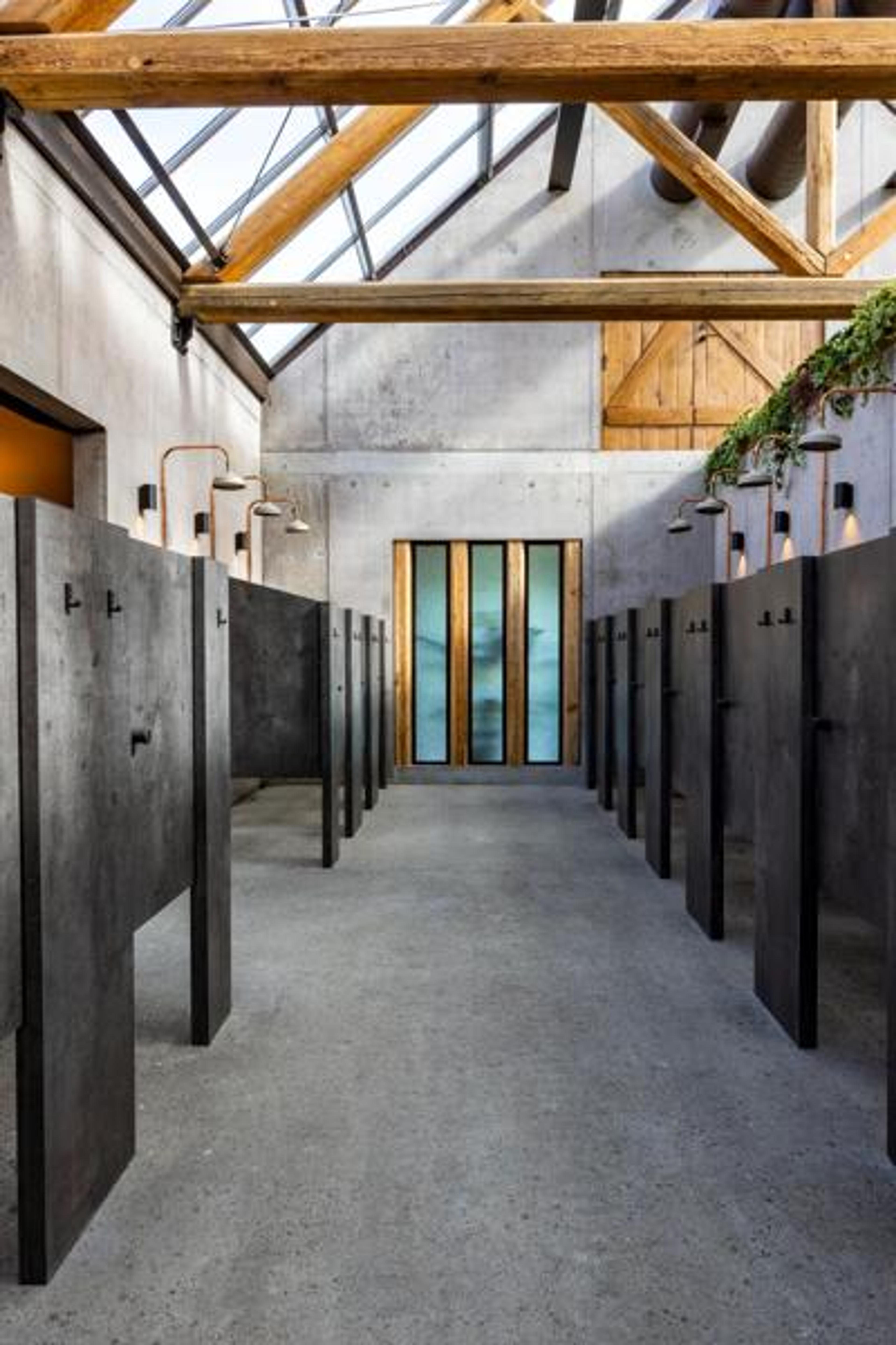 A modern industrial changing facility with concrete walls, exposed wooden beams, and skylights. Dark grey individual stalls line a central aisle, illuminated by industrial-style lights.
