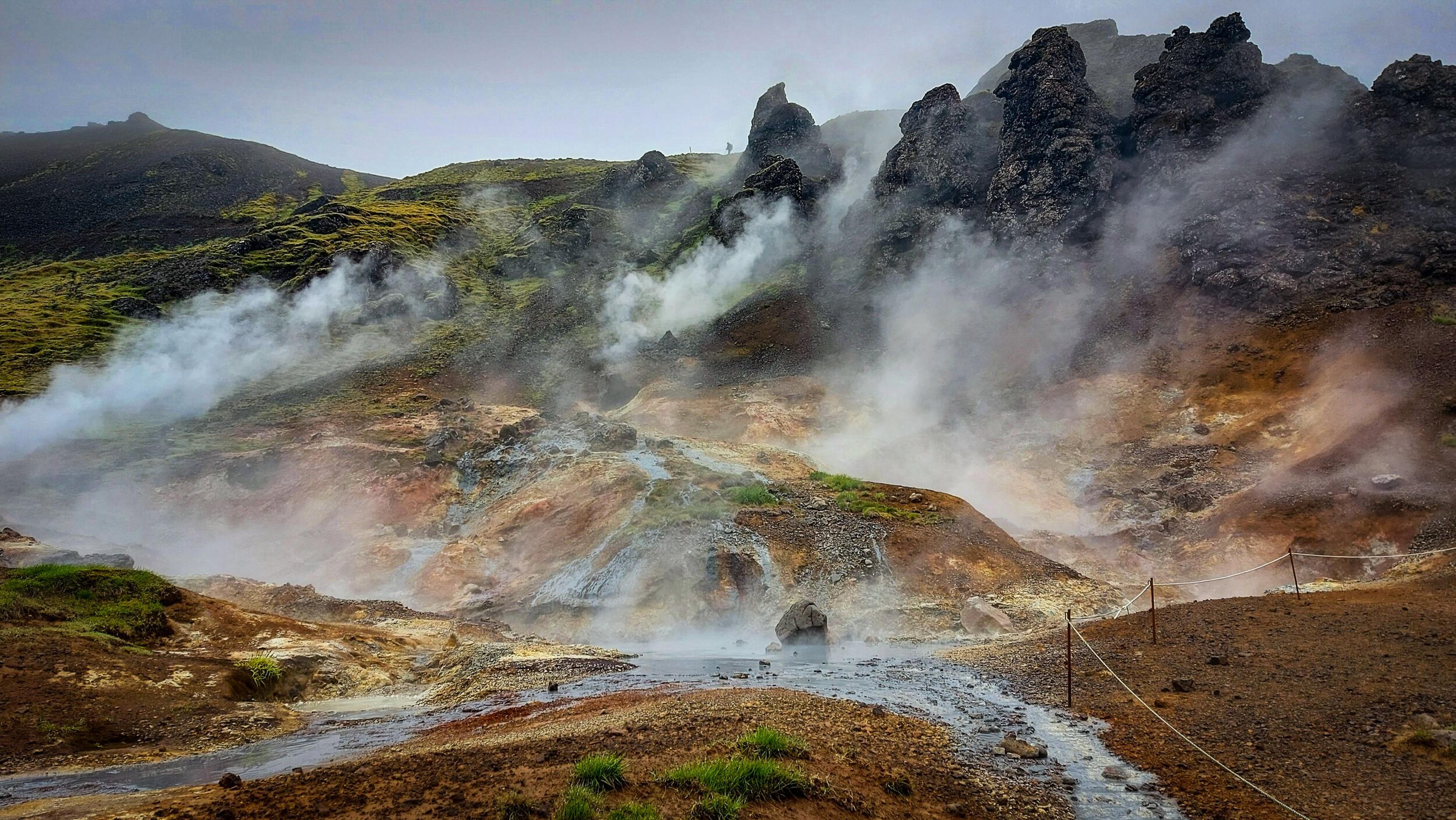 Steaming geothermal landscape with colorful mineral deposits, rocky hills, and a small stream.