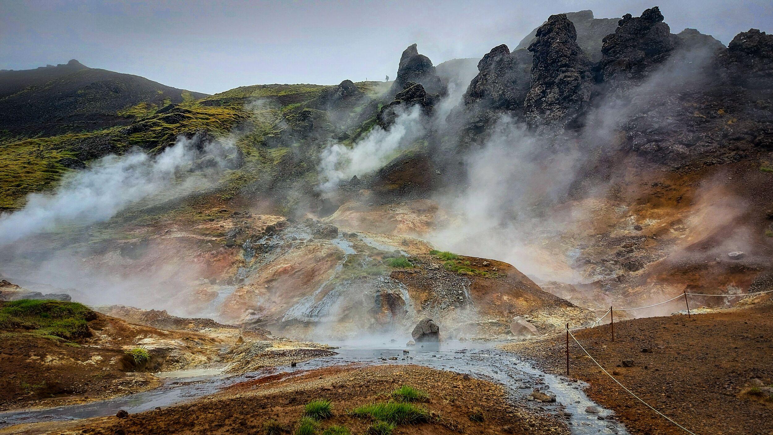 Steaming geothermal valley with colorful hills, dark rock formations, and a stream.