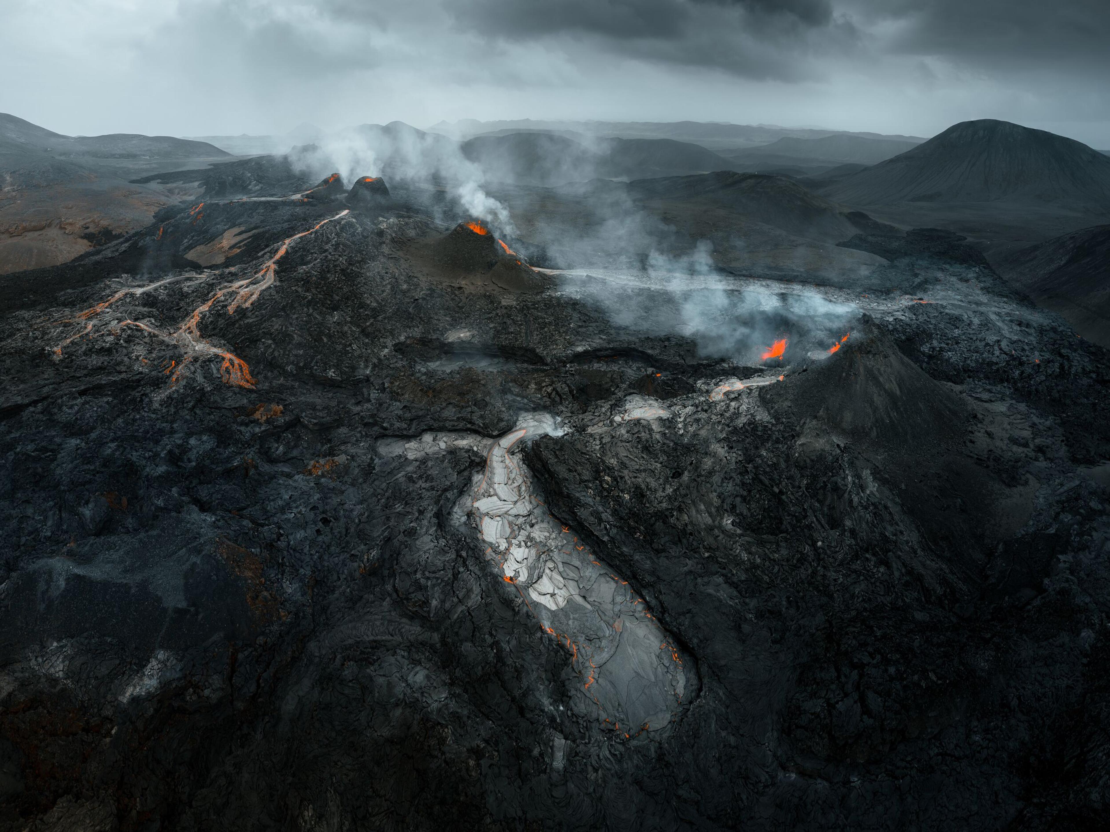 Aerial view of an active volcano with smoke and lava flows against a stormy sky.