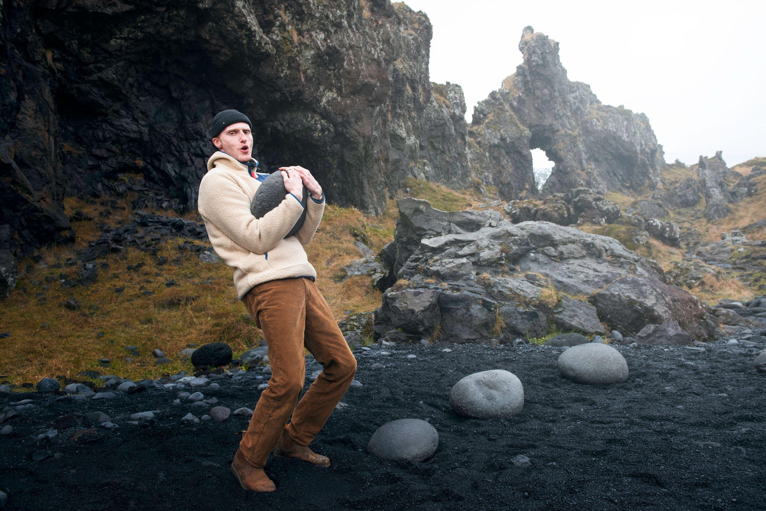 A man testing his strength by attempting to lift the traditional Icelandic lifting stones on Djúpalónssandur beach, with the rugged coastline and crashing waves as a backdrop.