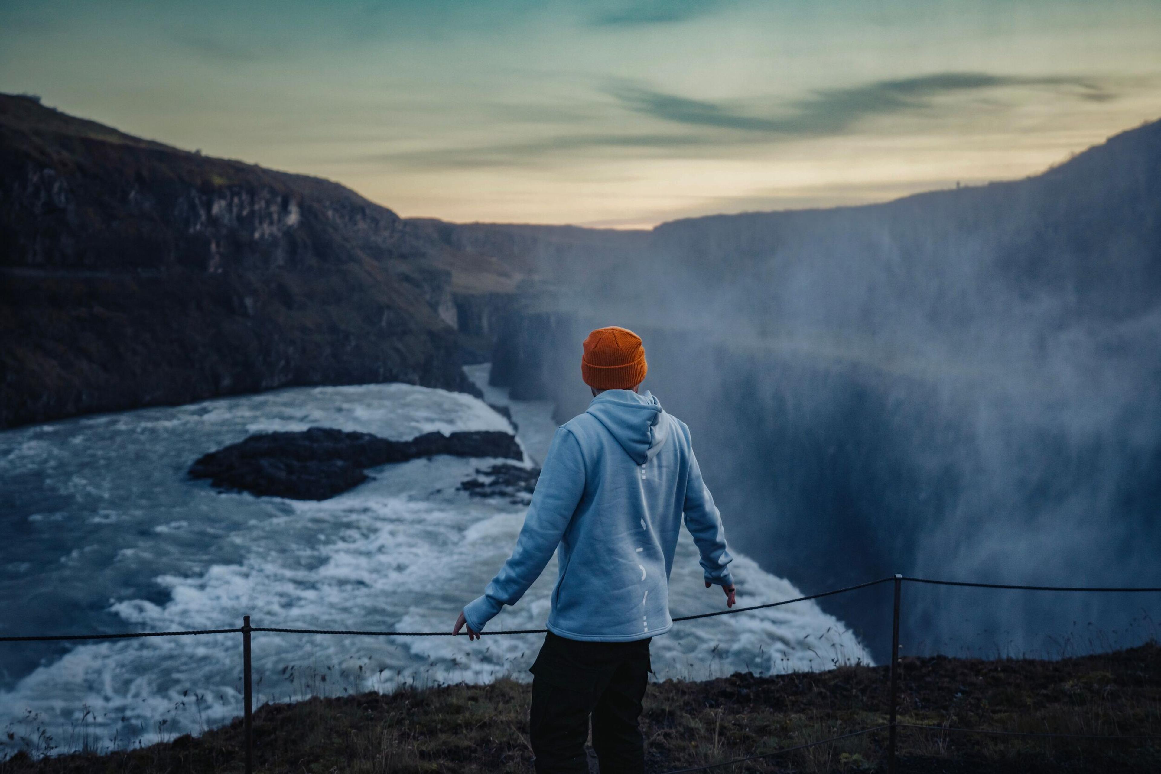  A person in a light blue hoodie and orange beanie stands near a dramatic waterfall, looking out over the rushing water and deep canyon at sunset. Mist rises around them, adding a moody atmosphere.