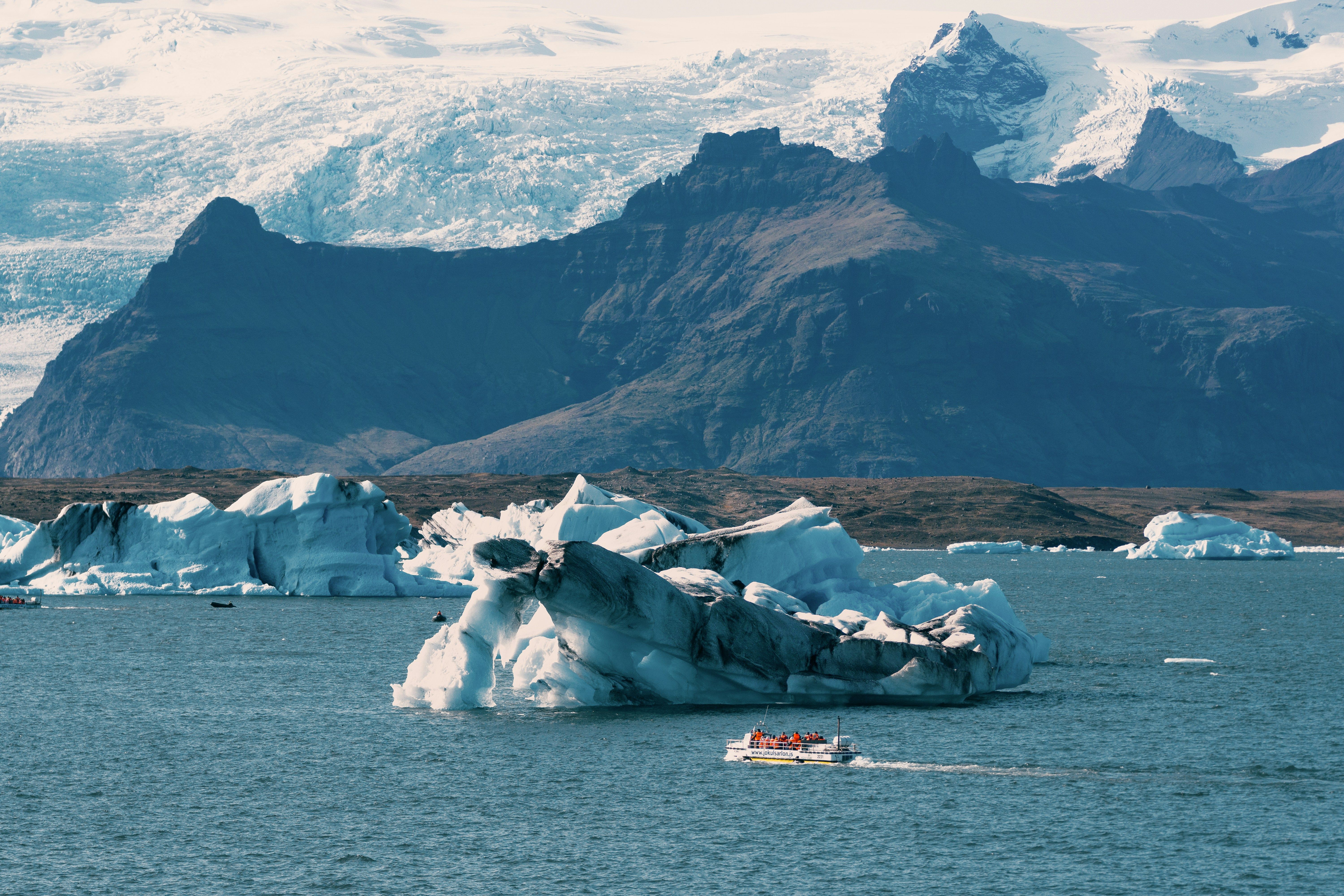 Boat tour navigating among large icebergs in a glacial lagoon with mountainous terrain and ice-covered peaks in the background in Iceland.