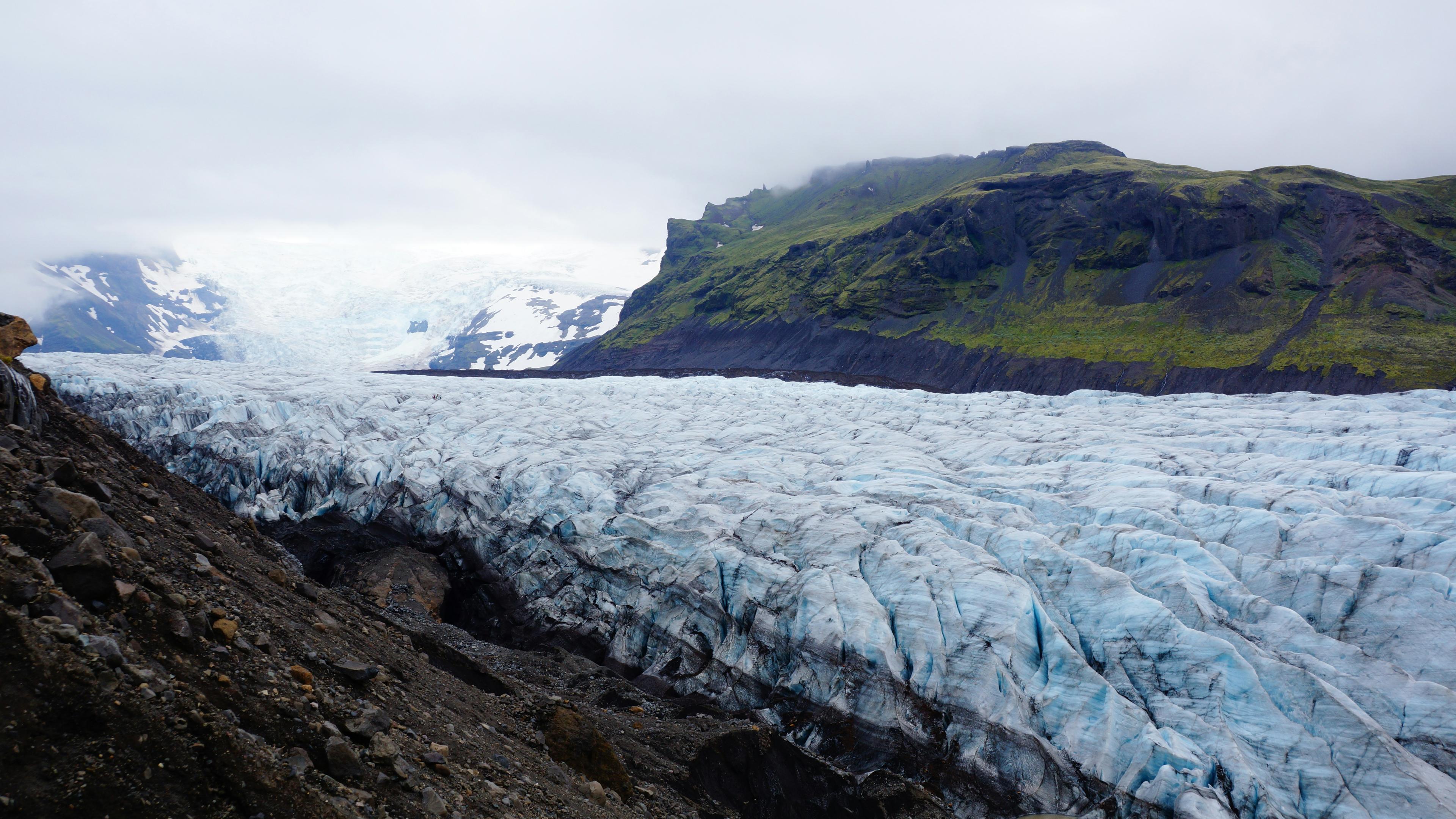 An expansive view of the Vatnajökull glacier in Iceland, showcasing its deep blue crevasses and rugged ice formations. The glacier is surrounded by contrasting green cliffs and dark volcanic rock under a cloudy sky, highlighting Iceland's dramatic and unique landscape.