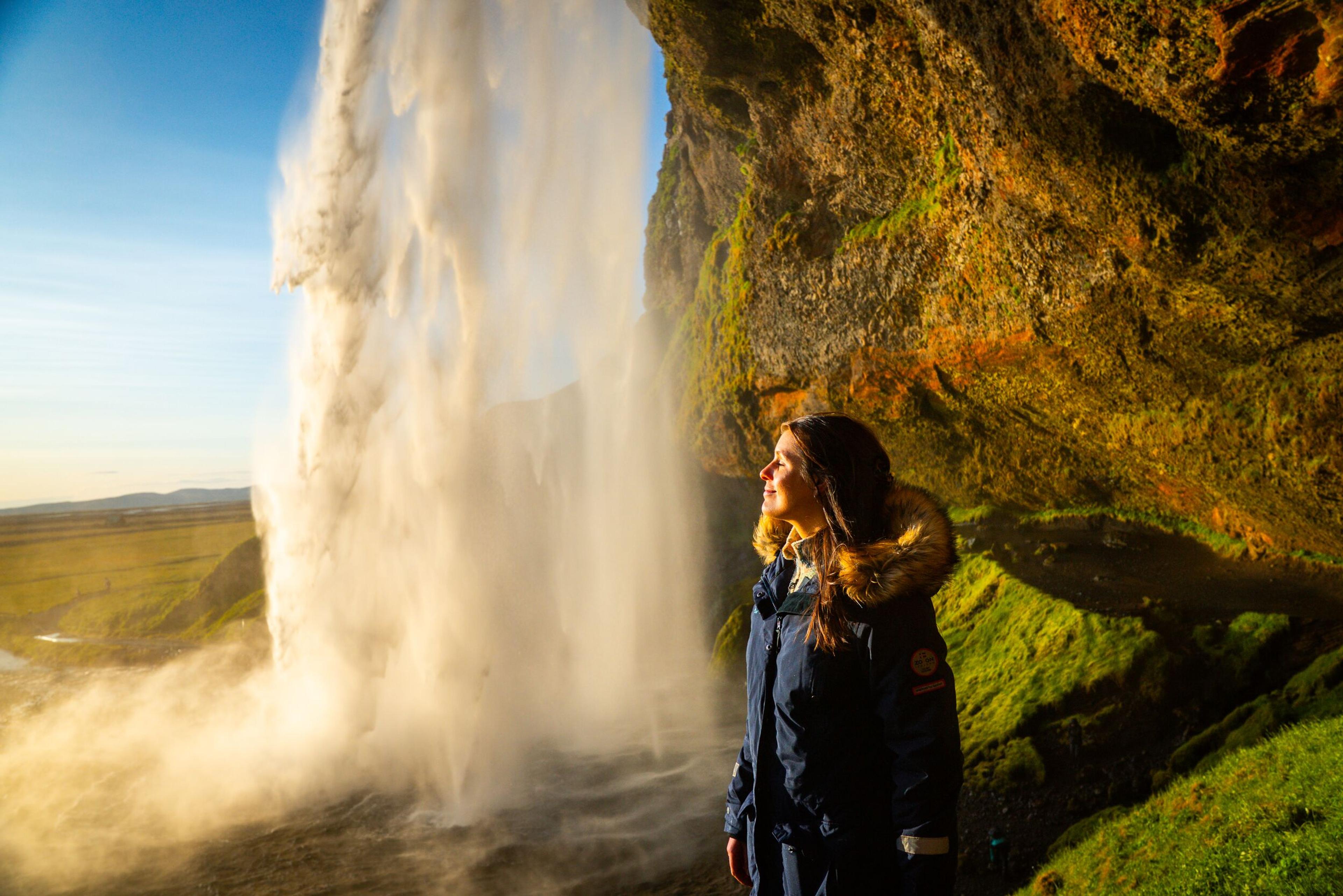 A woman in a warm jacket stands behind a sunlit waterfall next to a mossy cliff.