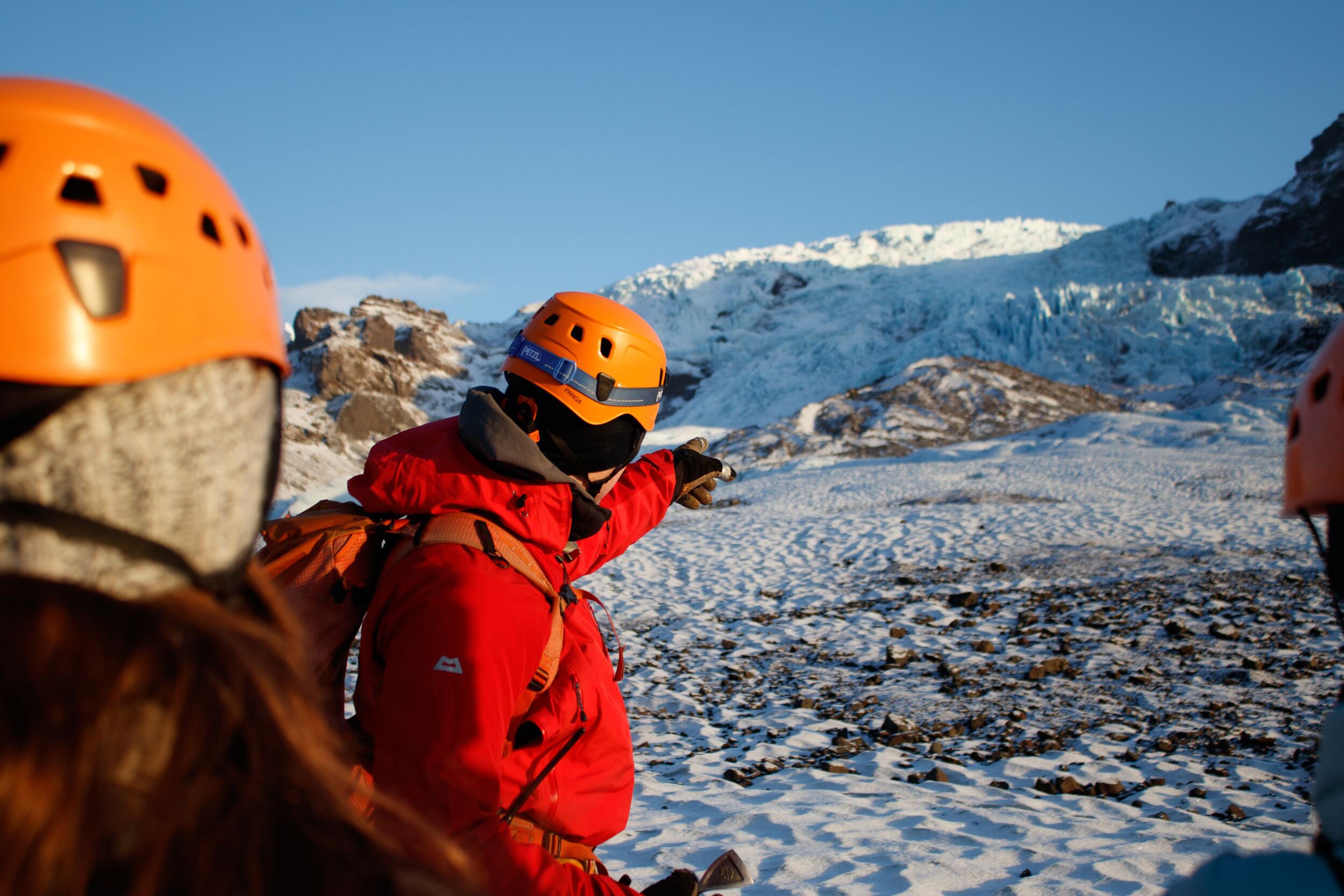 People in orange helmets on a snowy landscape, one pointing at a large glacier.