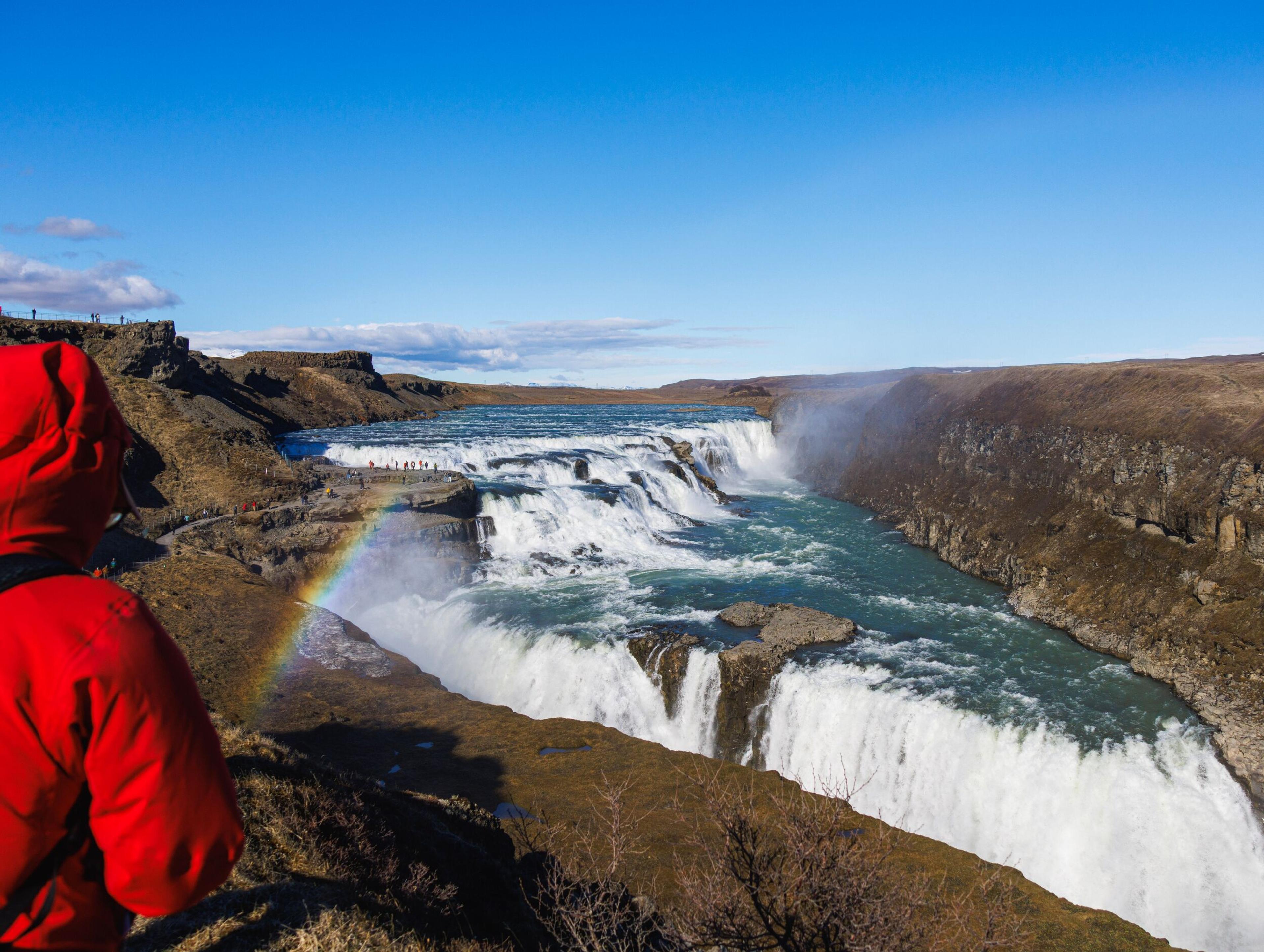 A person in a red jacket views a powerful waterfall with a rainbow appearing in the mist under a clear blue sky.