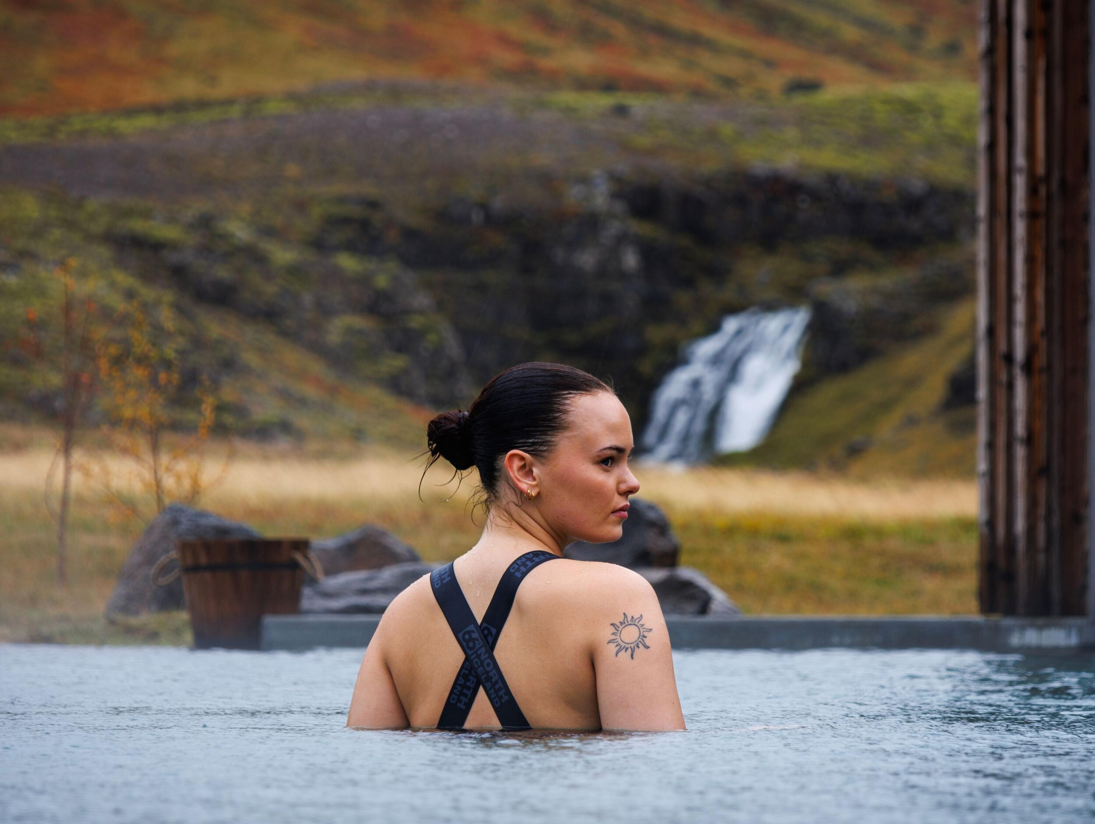 A woman with a sun tattoo on her shoulder relaxes in a geothermal pool with a waterfall and hills in the background.