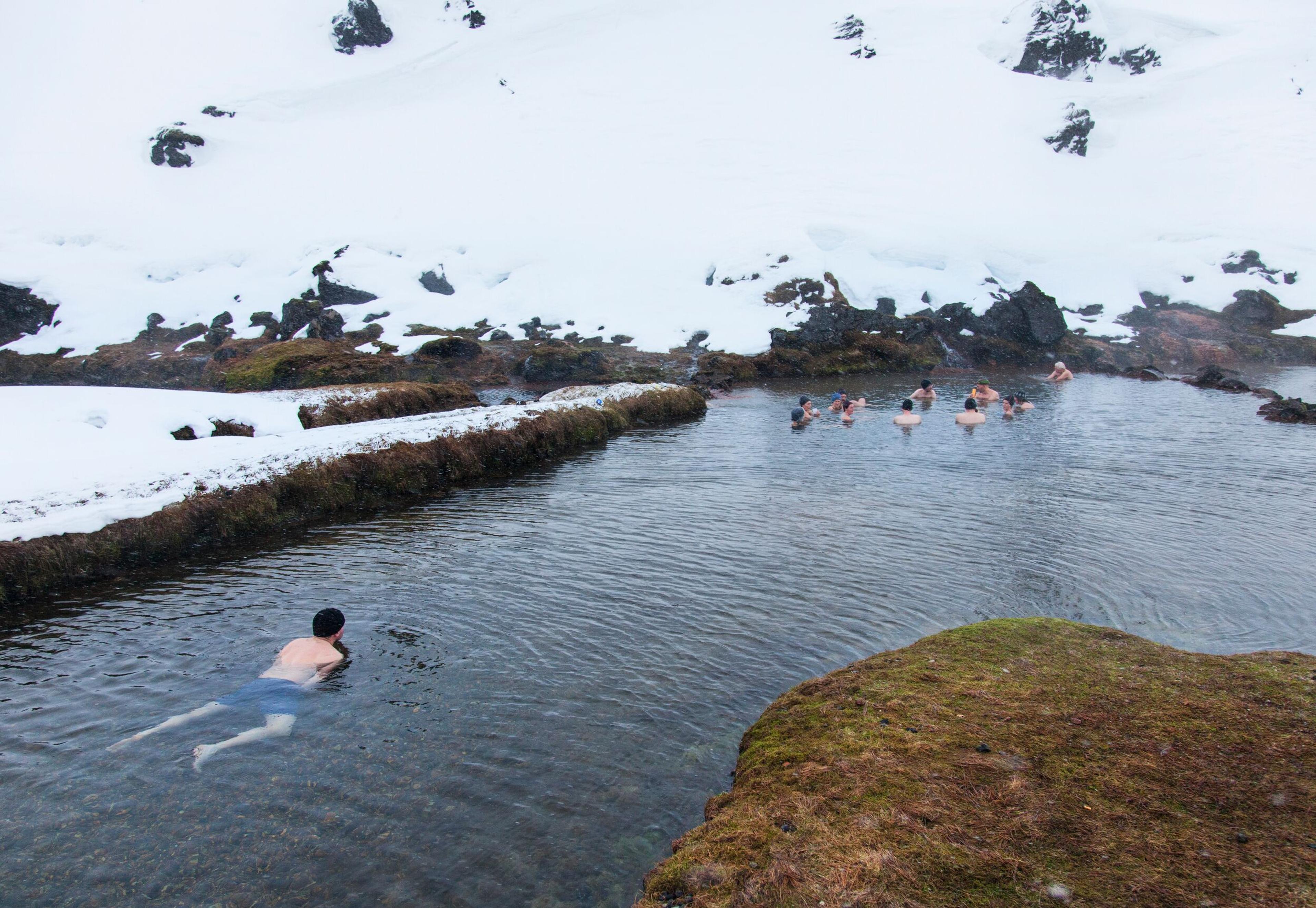 People swimming in a hot spring surrounded by snow in Landmannalaugar during winter, with one person swimming alone in the foreground.