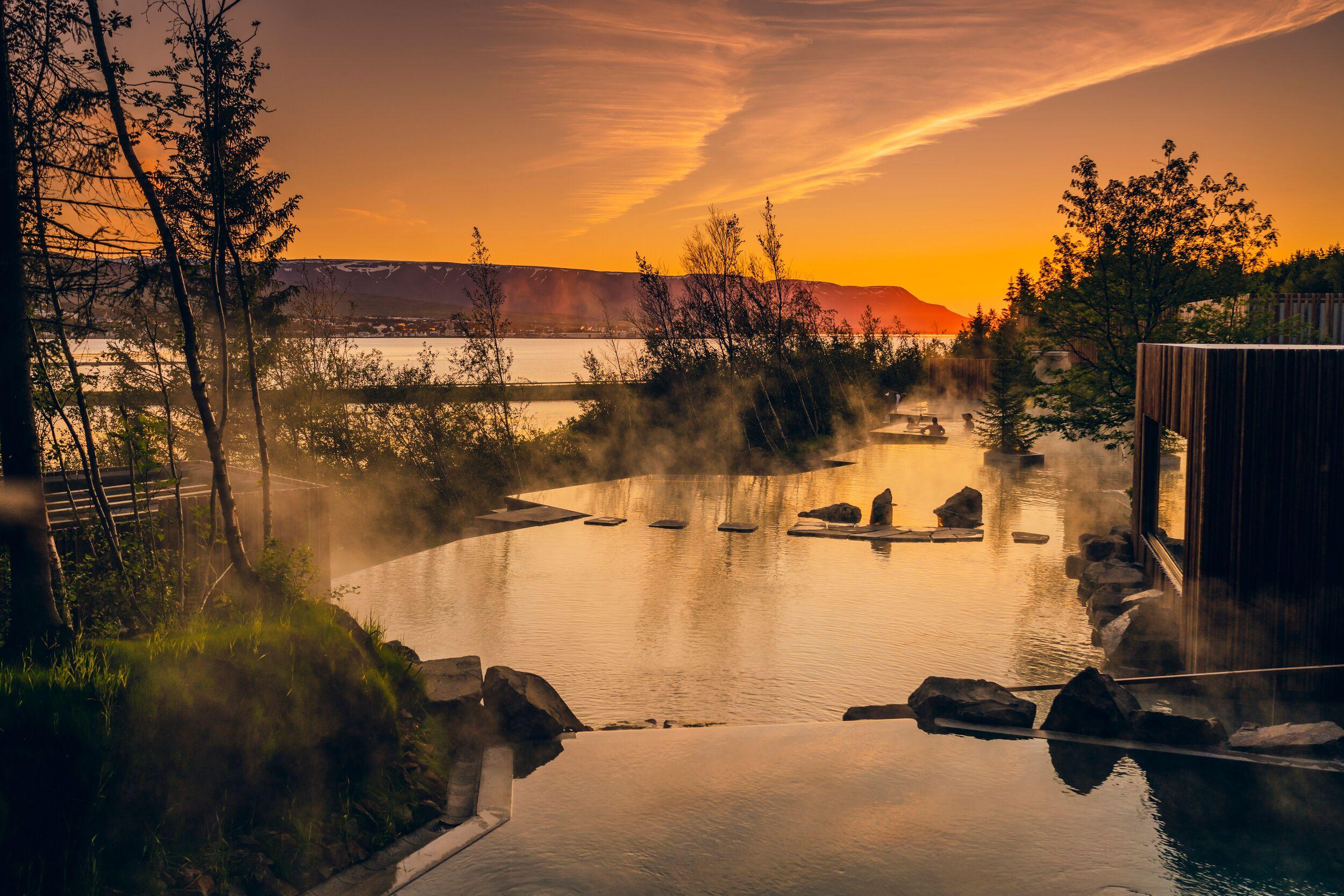 Steaming geothermal pools reflecting a golden sunset, with a lake and mountains in the distance.