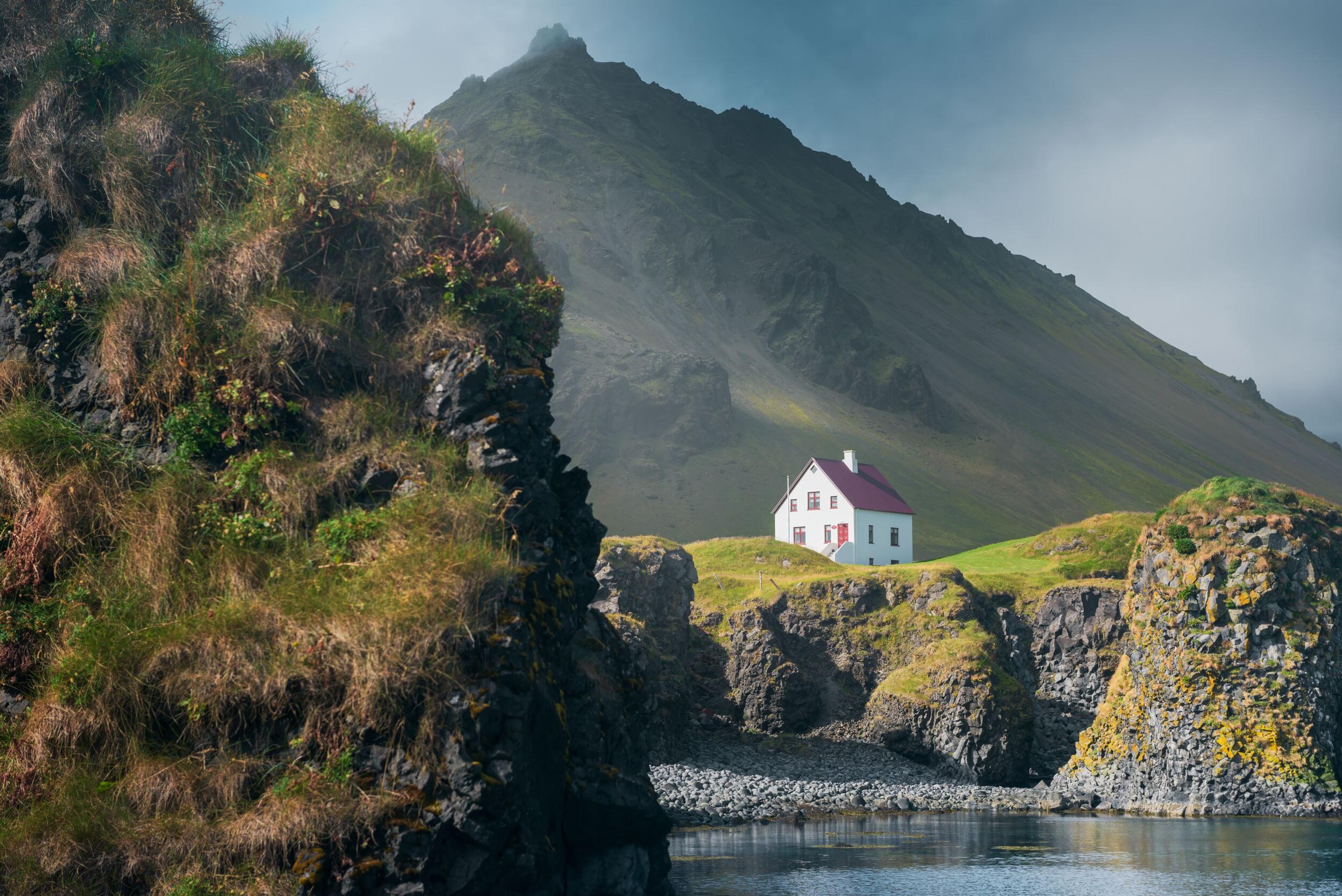 A solitary white house with a red roof stands in stark contrast to its surroundings, nestled among green cliffs and rocky shores near a tranquil body of water, with a towering mountain in the background.