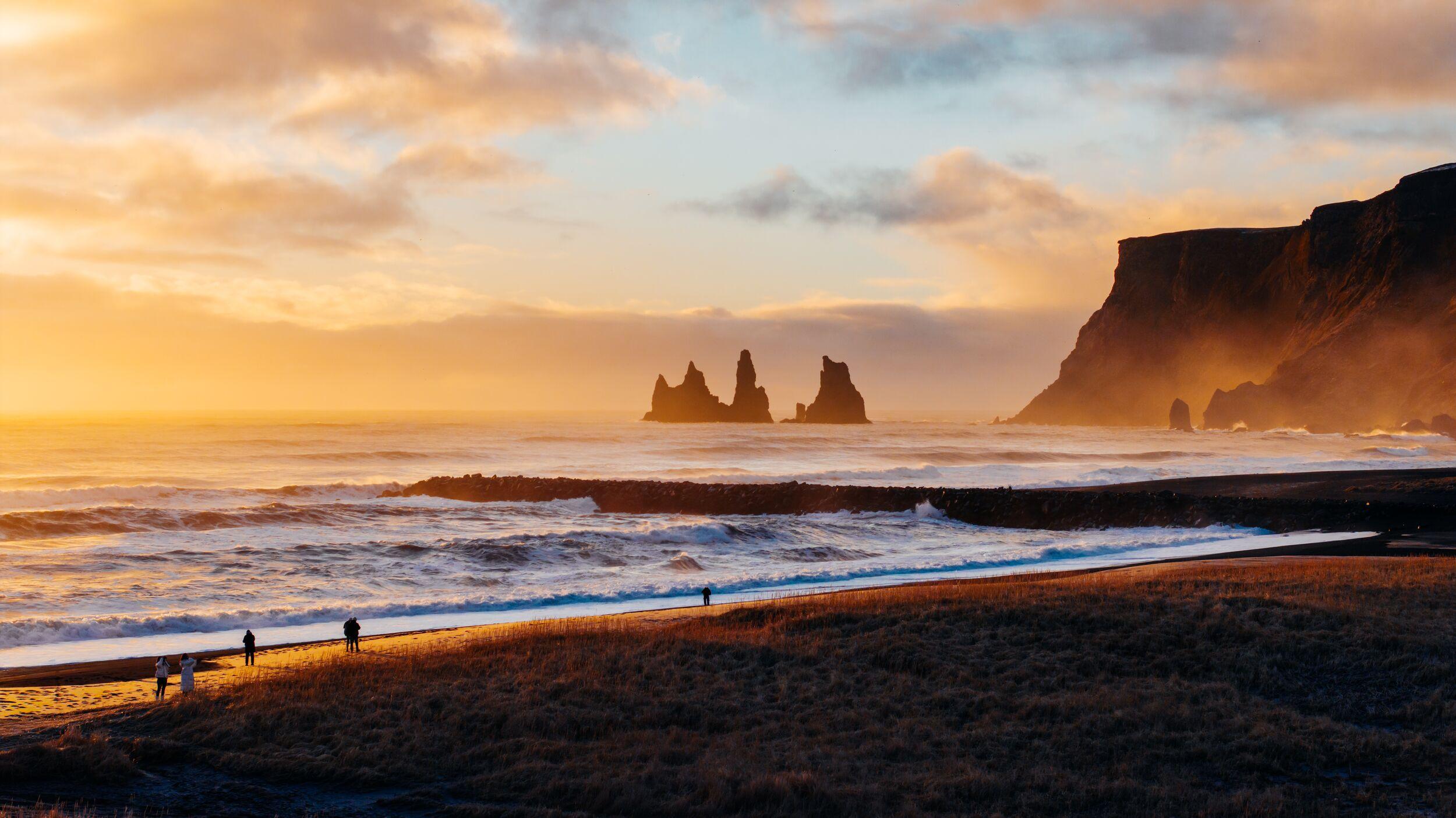 Golden sunset over a black sand beach with crashing waves, sea stacks, a dark cliff, and distant people.