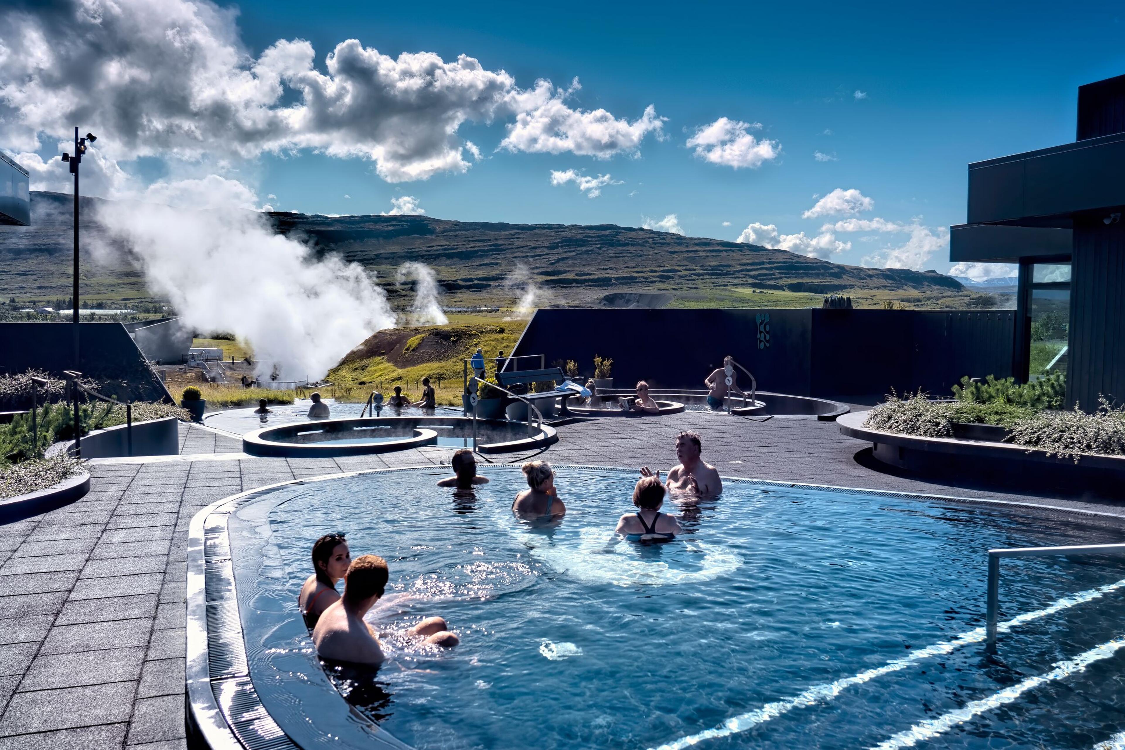 The Krauma geothermal baths in Iceland, featuring circular hot tubs surrounded by modern black stone design and a backdrop of steaming geothermal vents and grassy hills.