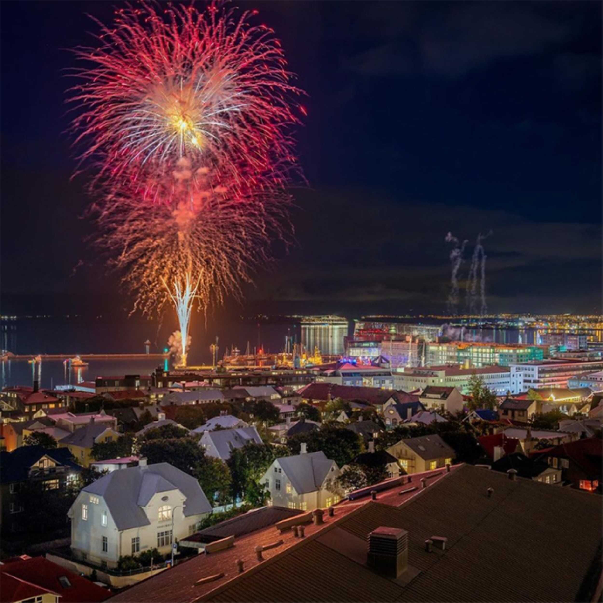 Colorful fireworks light up the night sky over Reykjavík's harbor, with city lights and residential rooftops in the foreground during Culture Night.