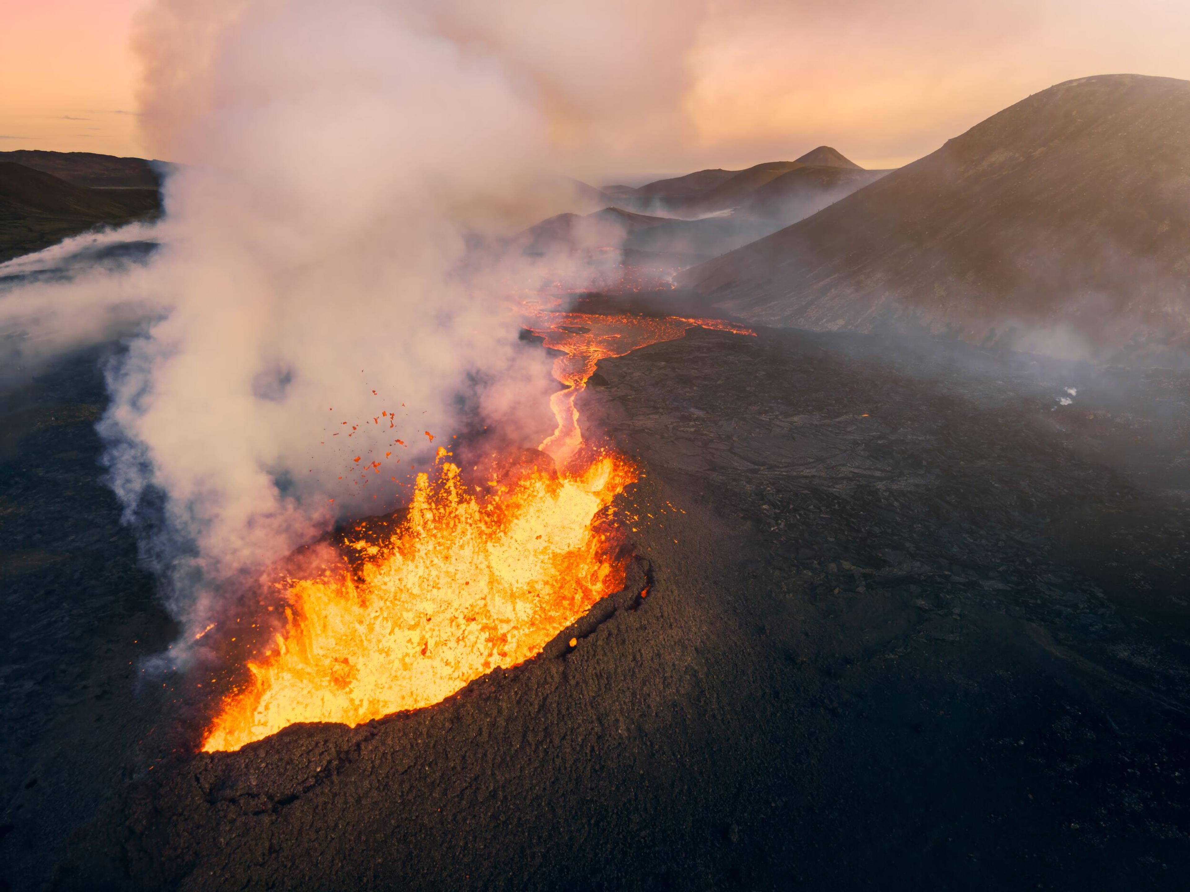 A fiery volcanic eruption with bright orange lava flowing and smoke rising against a backdrop of mountainous terrain during sunset.