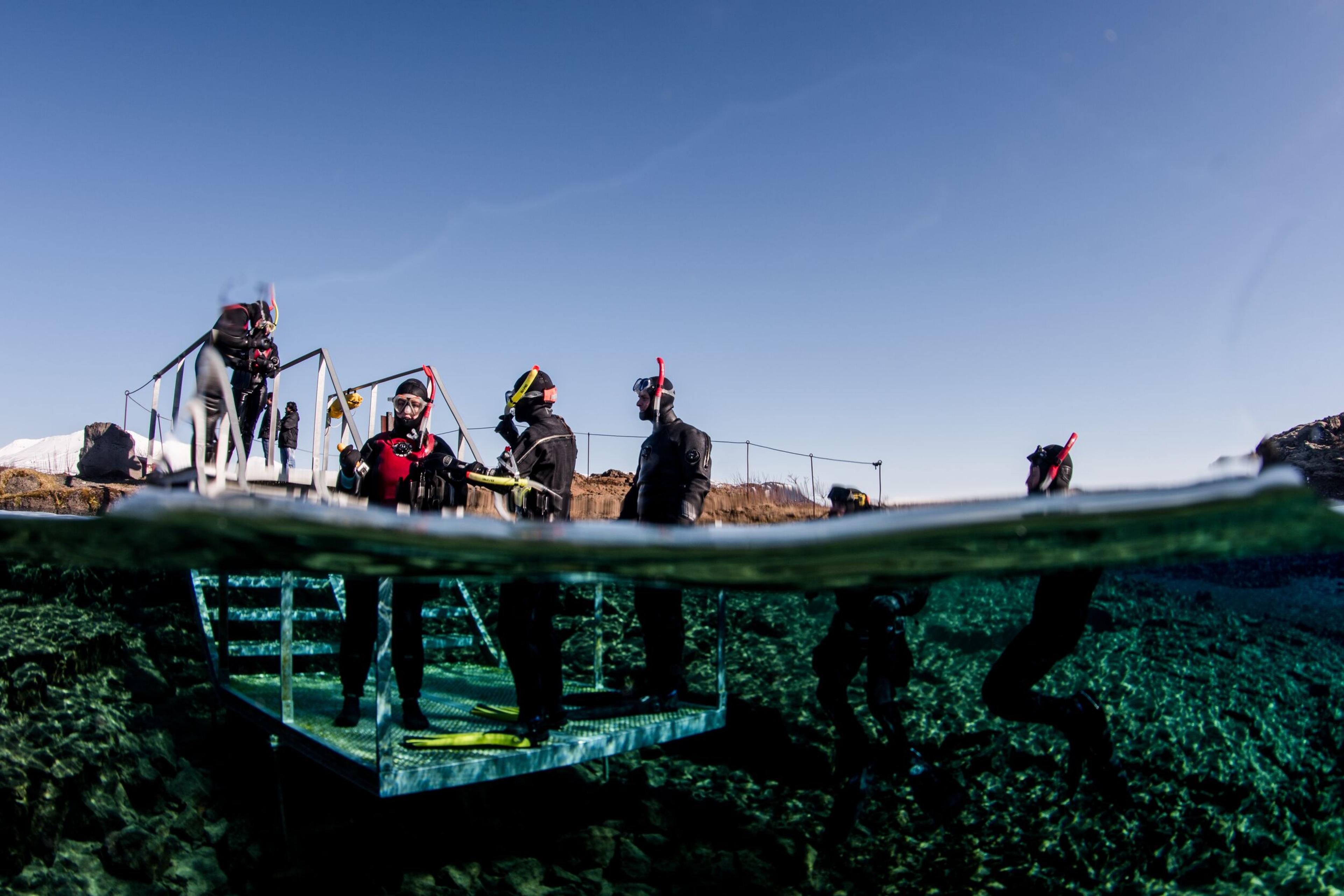 Split view of snorkelers in wetsuits on an entry platform and in incredibly clear blue water with a rocky bottom, under a sunny sky.