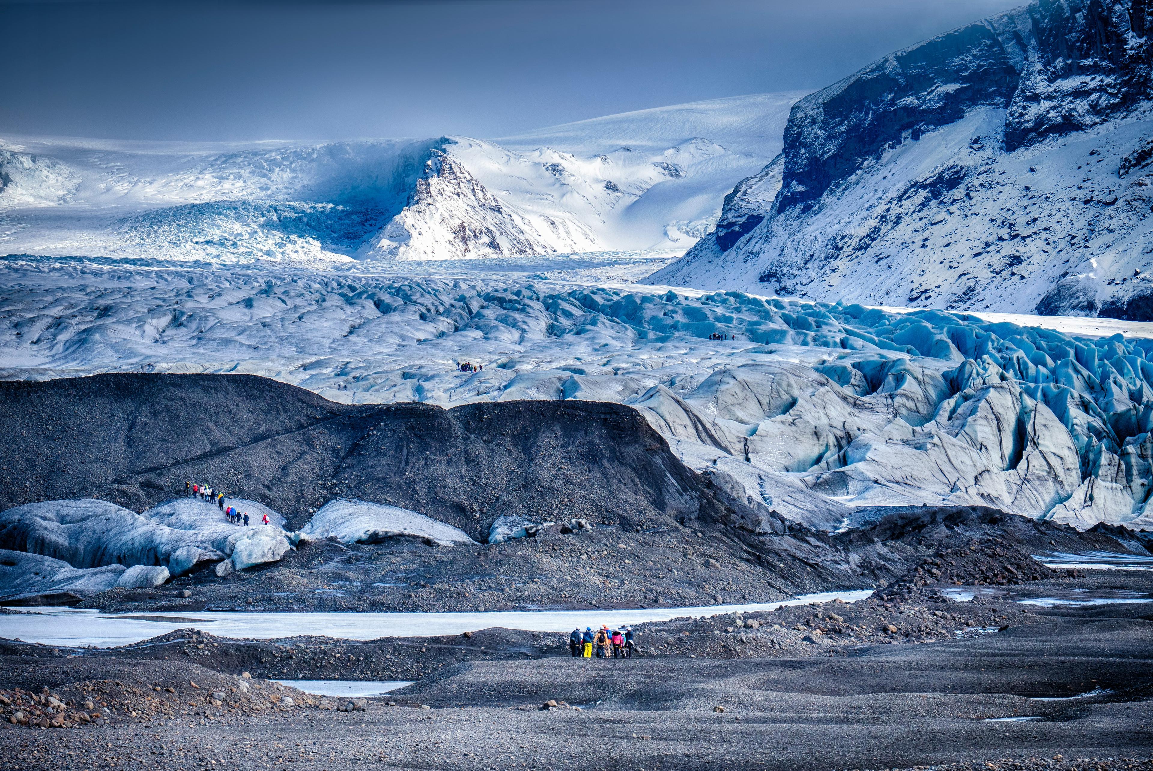 Groups of tourists near the edge of a vast glacier with deep blue crevasses and rugged ice formations, set against a backdrop of snowy mountains.