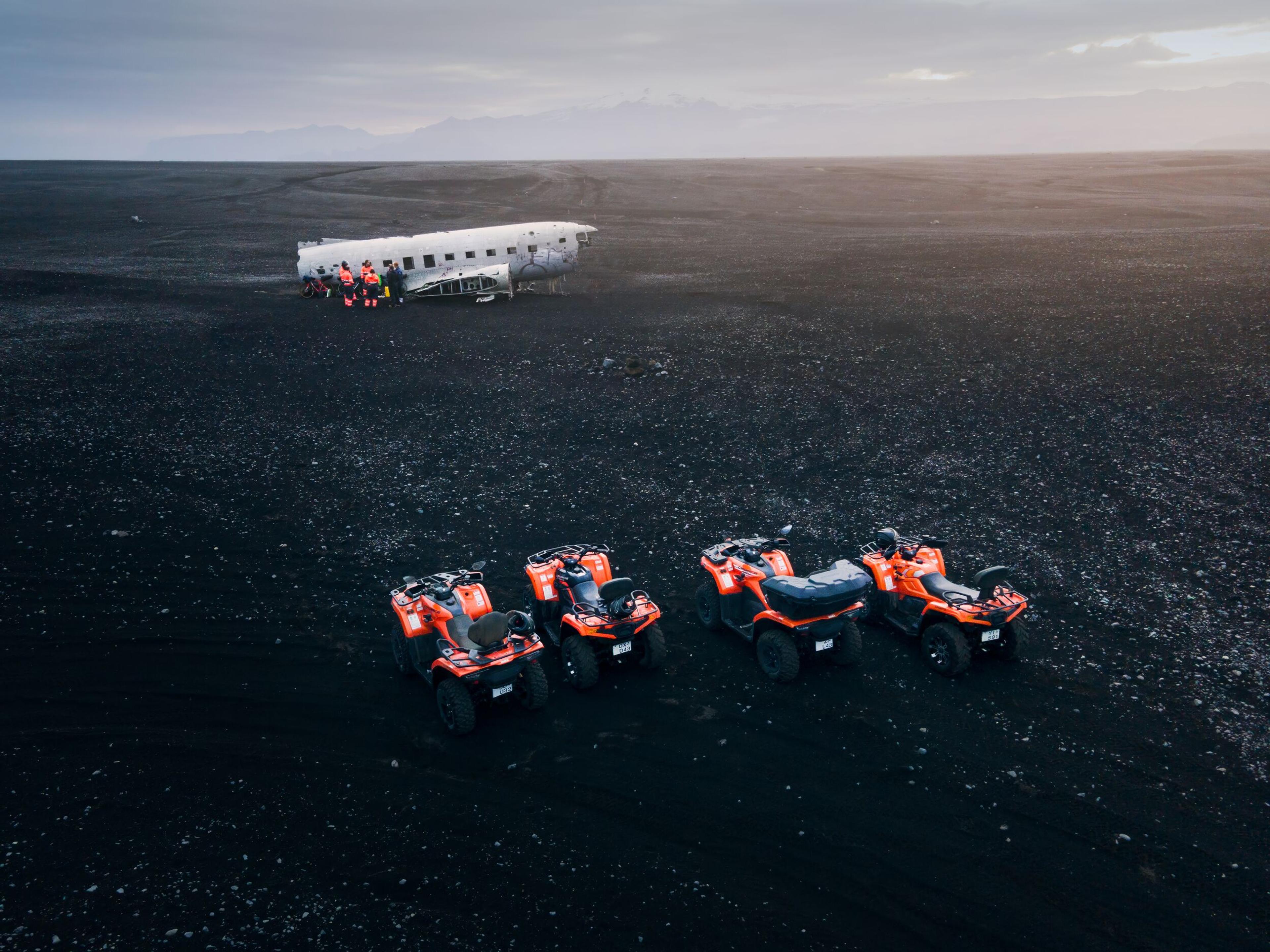 Adventurers gather around a historic plane wreck on Iceland's vast black sand expanse, with ATVs poised for the next thrill.