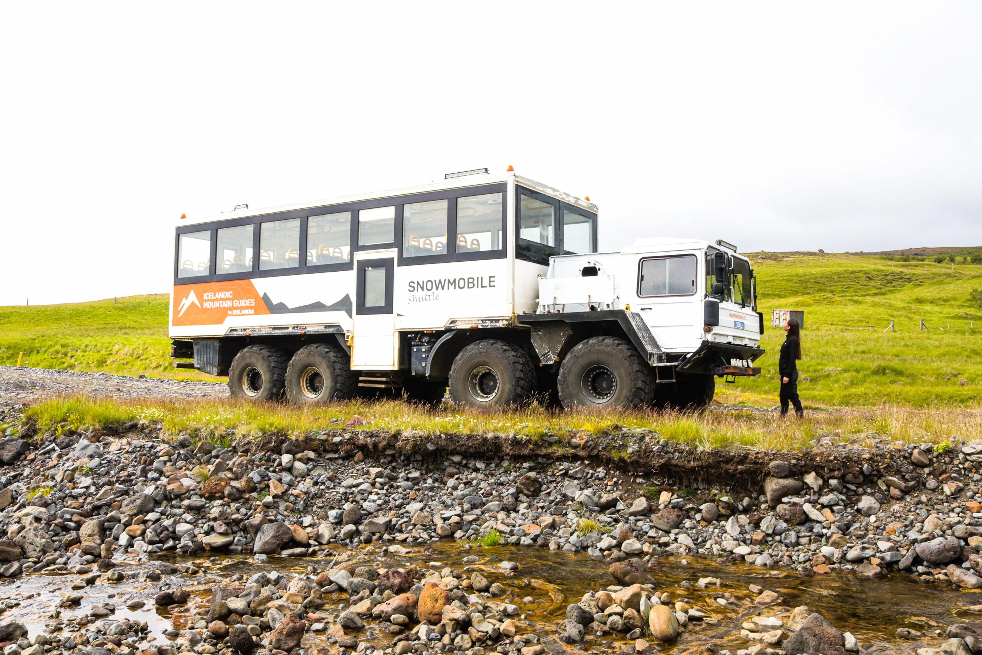 Large off-road mountain truck used for snowmobile shuttles, parked on rocky terrain with a person standing beside it.