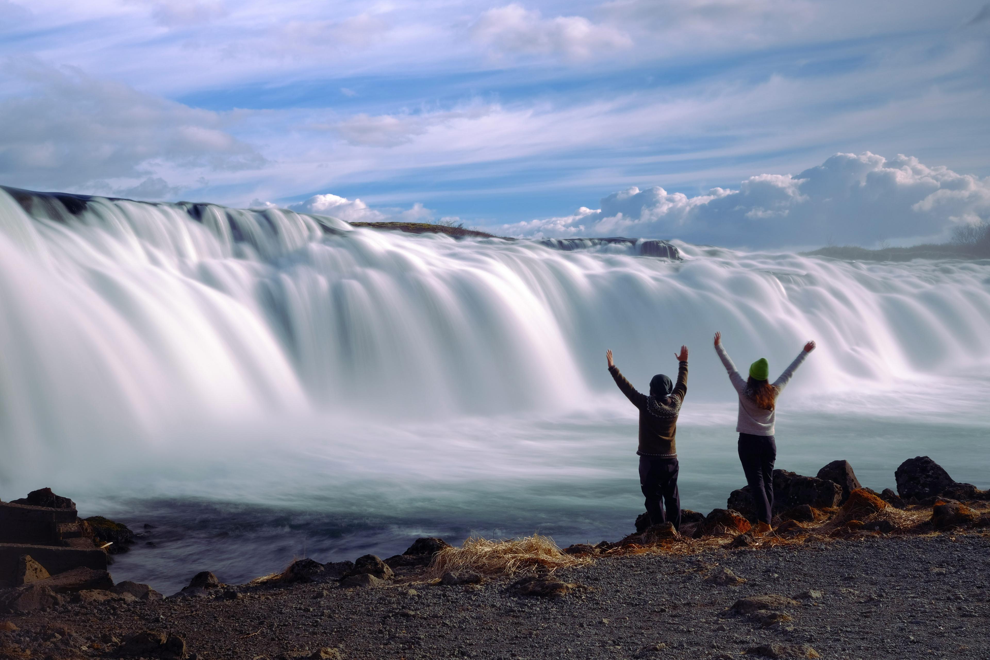 Two people with raised arms stand in front of a majestic, wide waterfall, their silhouettes expressing joy and freedom against the powerful and beautiful cascade under a cloudy sky.