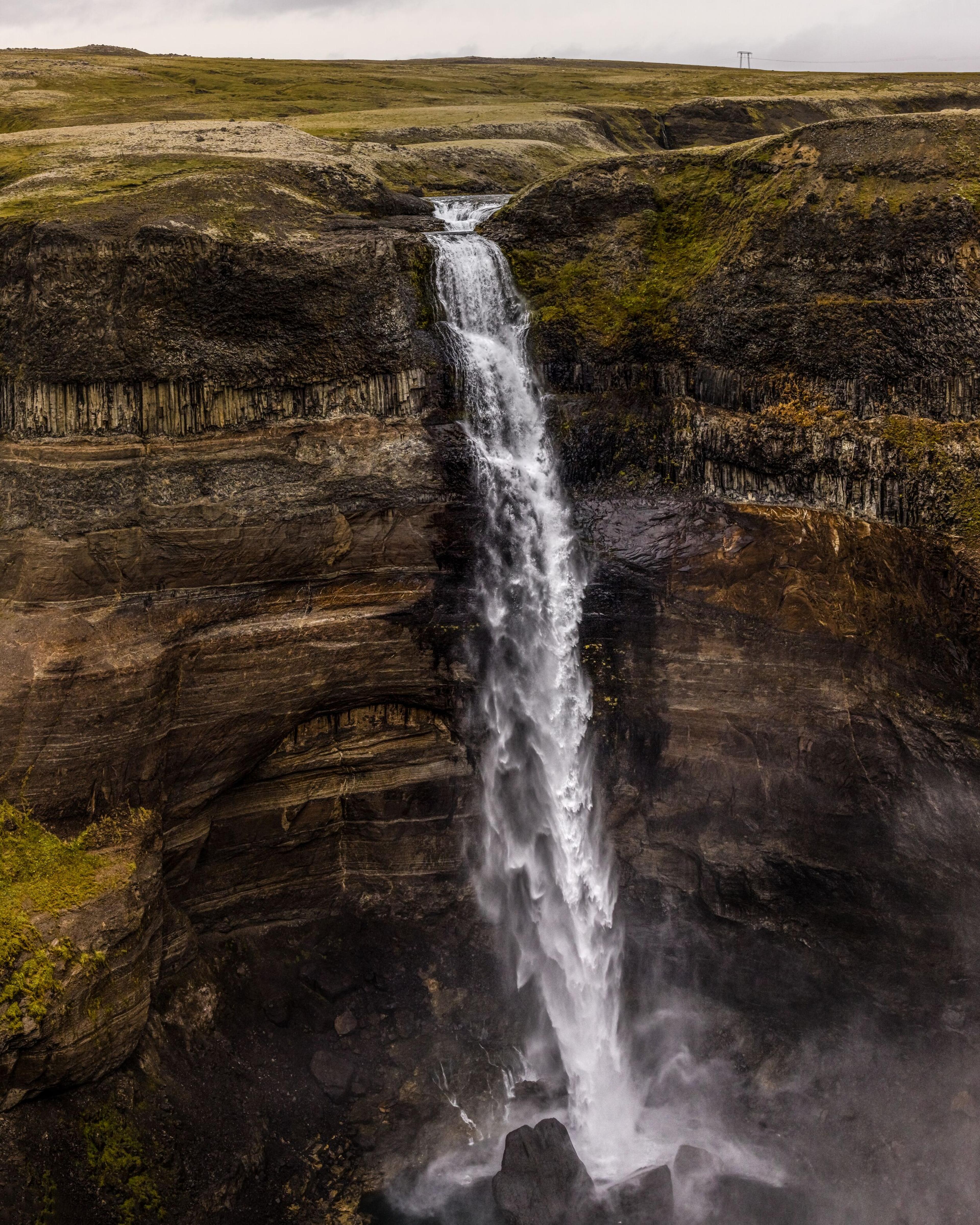 Tall waterfall cascading down a dark cliff with prominent basalt columns and green vegetation under a cloudy sky.