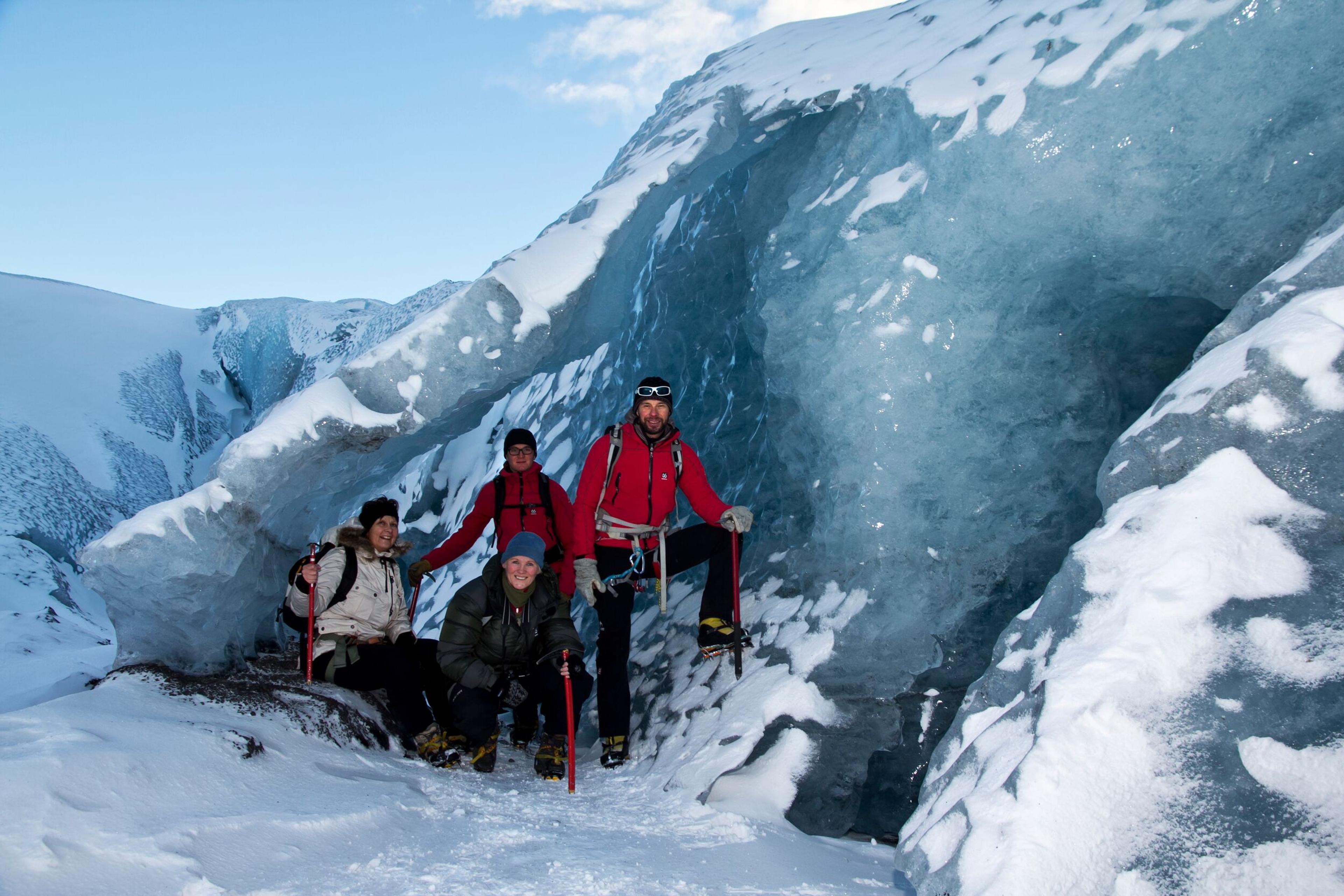 Group of four climbers posing at the entrance of an ice cave on Mýrdalsjökull glacier, surrounded by snow and ice formations.