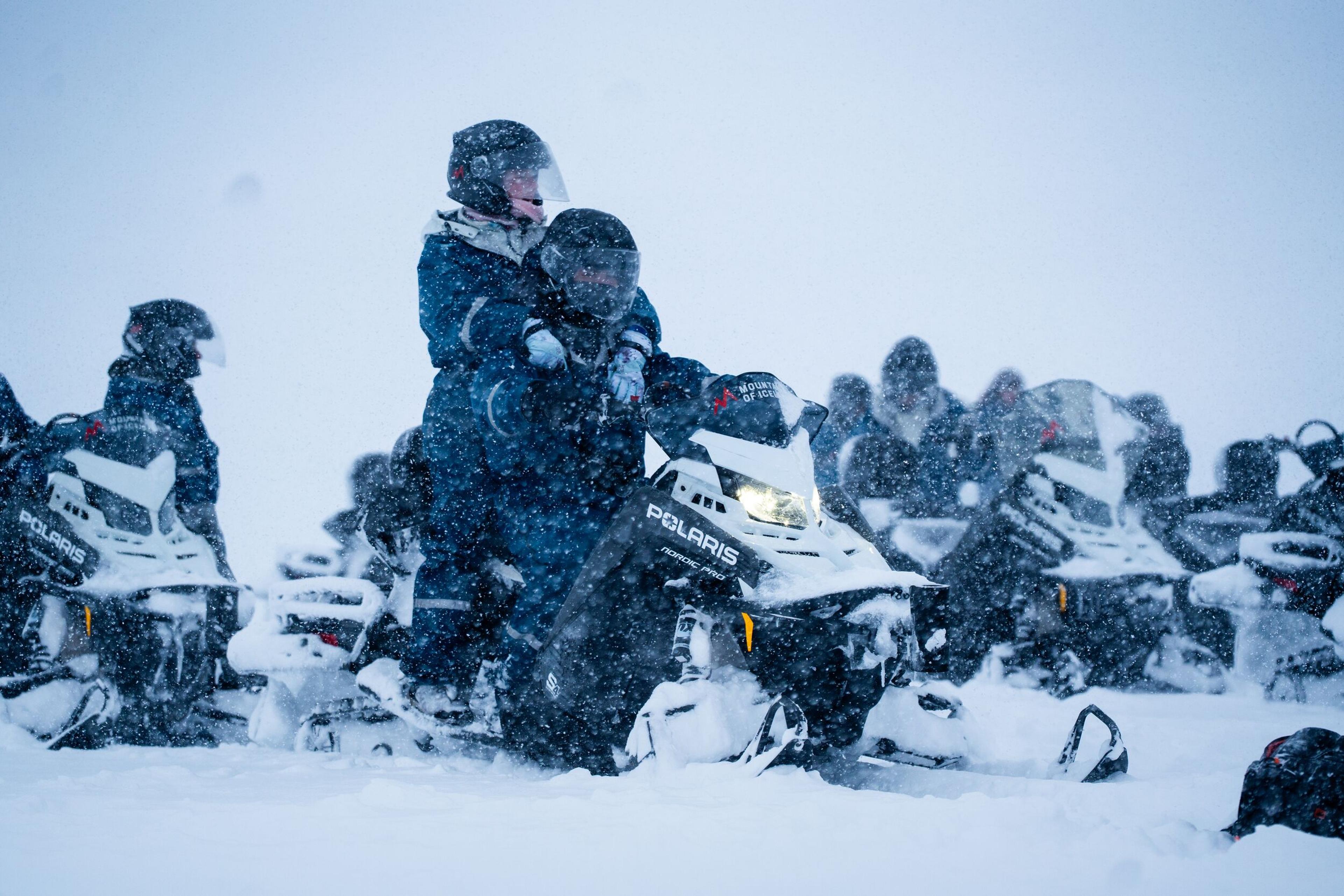 Two people on a snowmobile with a group of other snowmobilers in a heavy snowstorm.