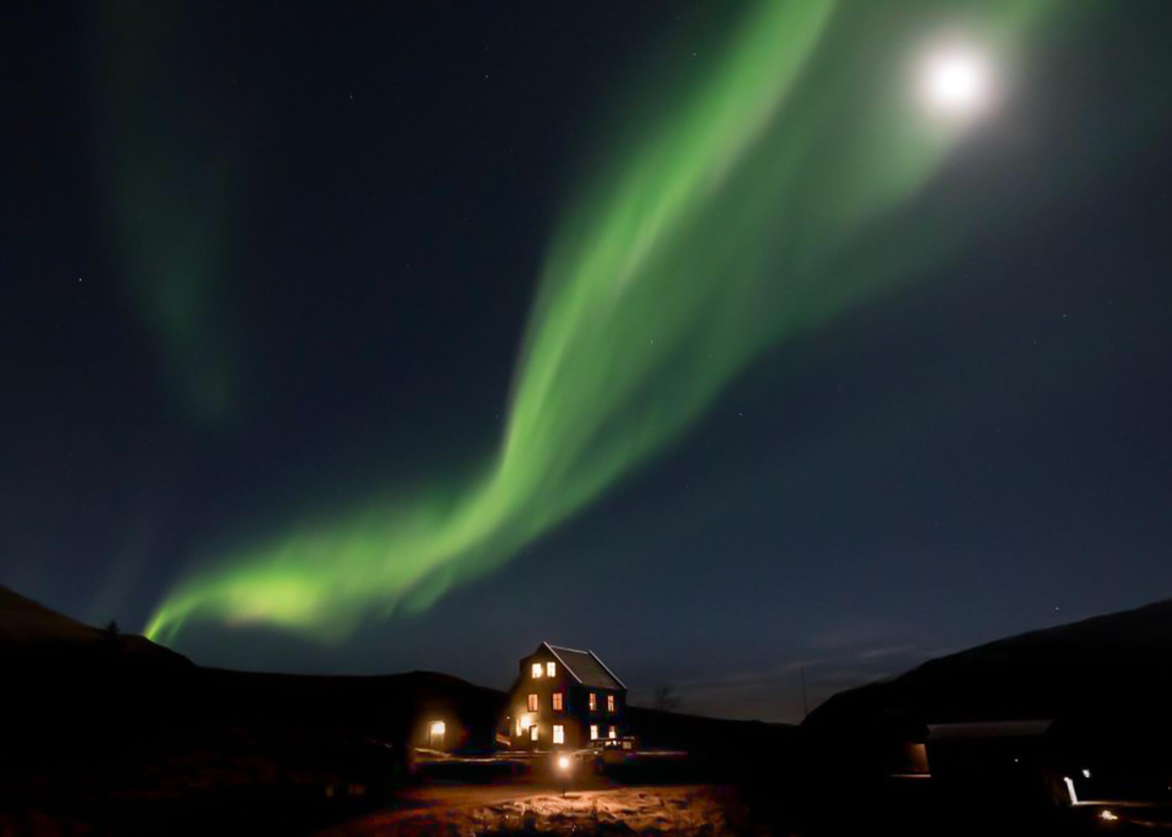 A solitary house stands under the night sky, bathed in the ethereal glow of the Northern Lights, their vibrant dance casting an otherworldly luminescence over the quiet landscape.