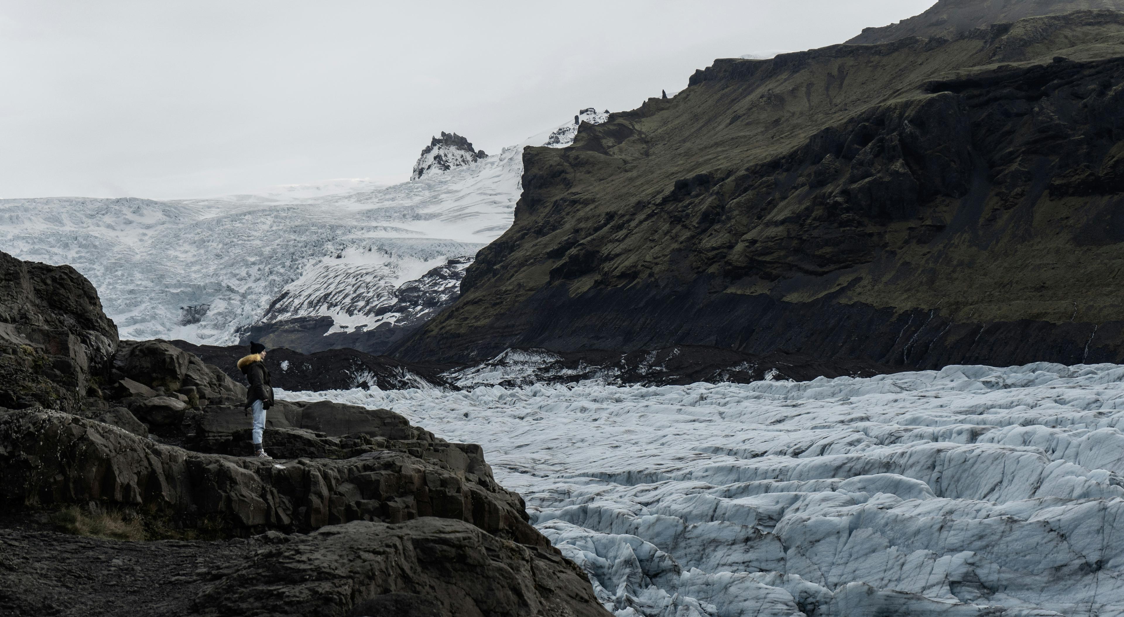 Traveler standing on rocky terrain, overlooking a vast glacier surrounded by rugged mountains and snow in Iceland.