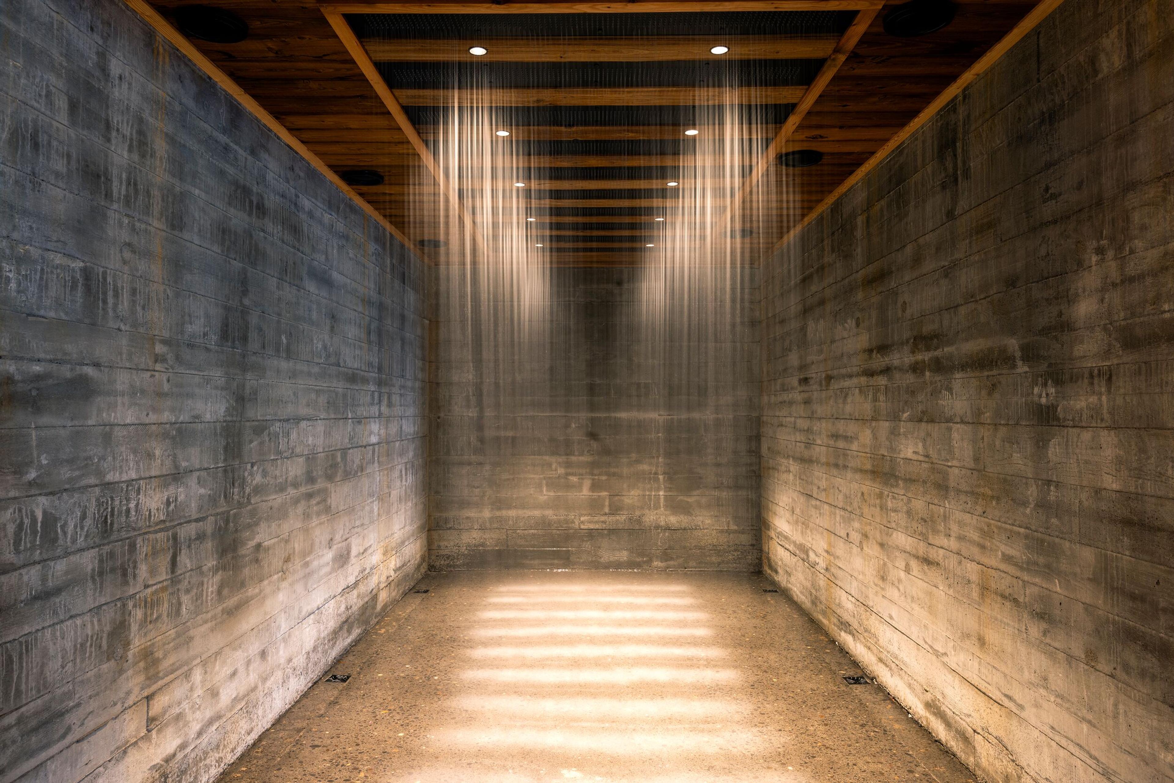 A narrow concrete room with a wooden beamed ceiling and fine vertical light streams casting striped patterns on the floor.