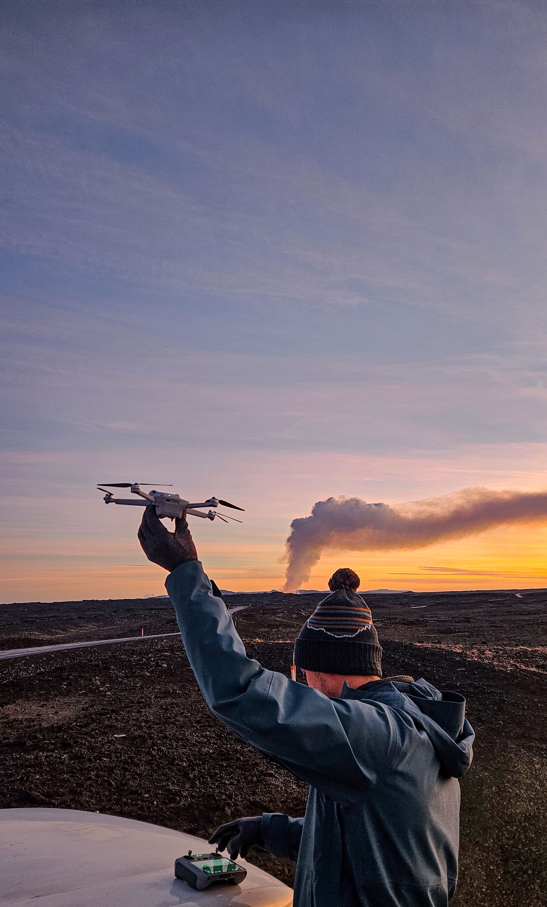 Person holding a drone against a sunset sky with a smoking volcano in the distance.