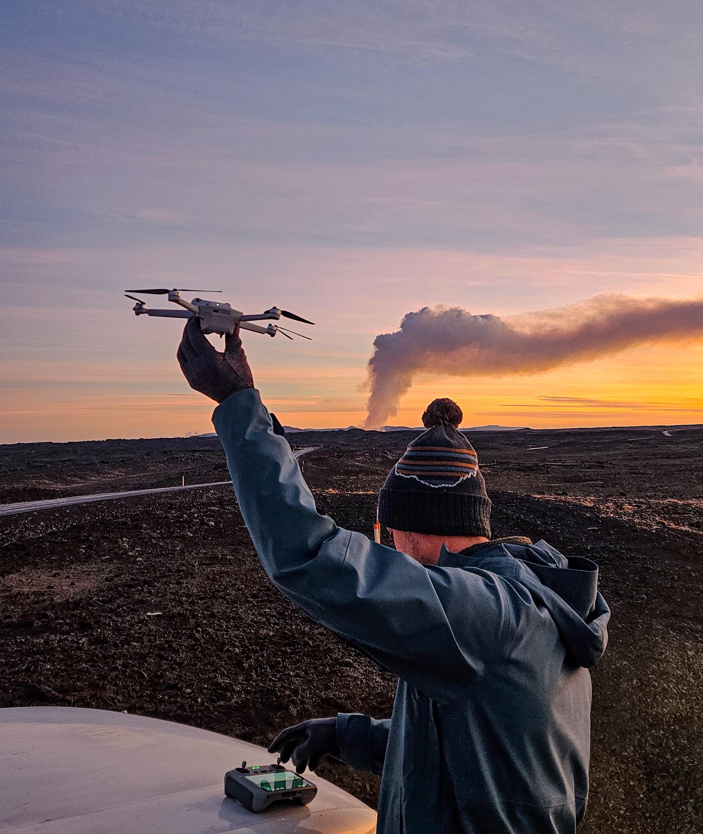 Person holding a drone against a sunset sky with a smoking volcano in the distance.