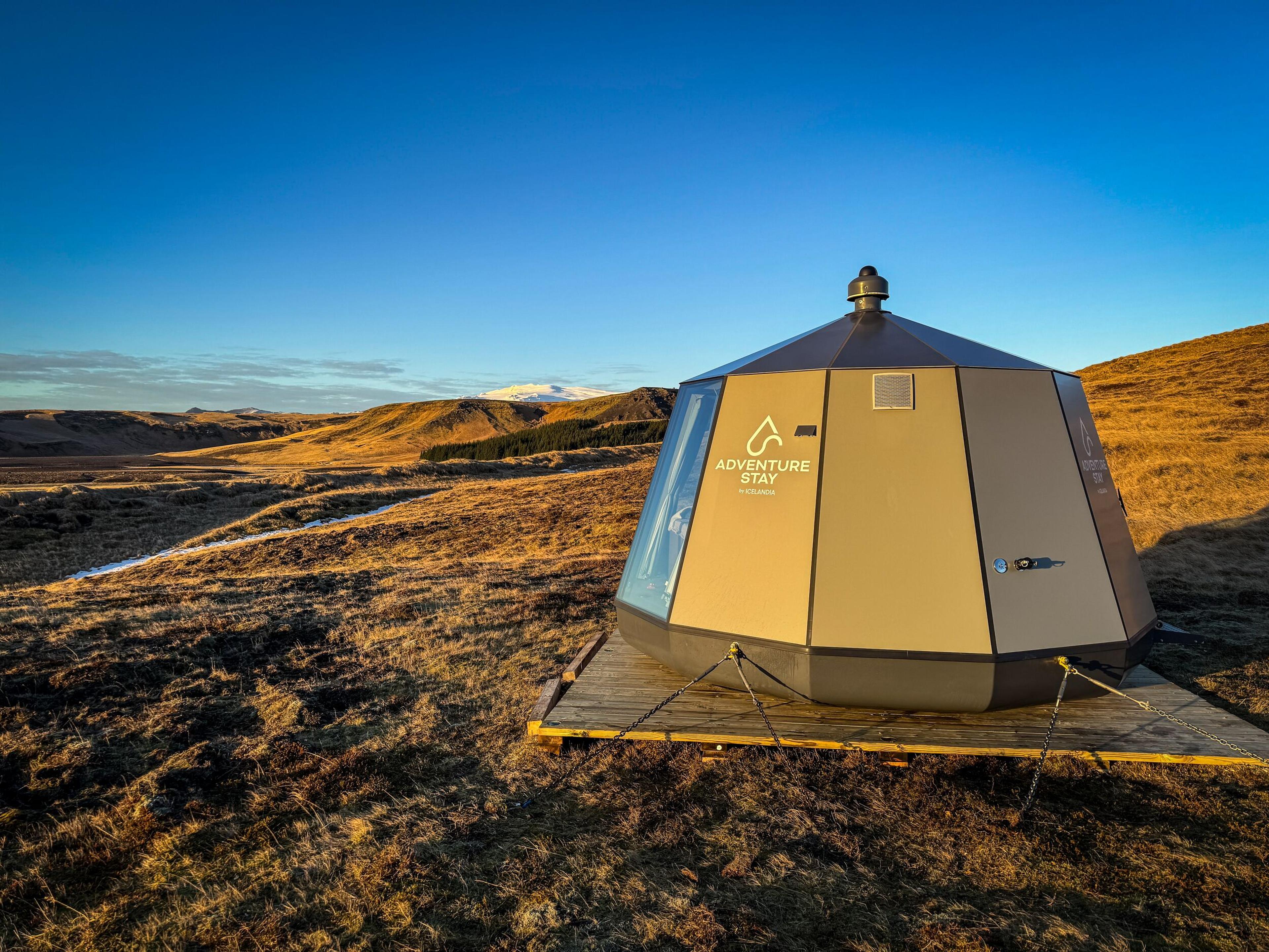 Modern octagonal 'ADVENTURE STAY' cabin with a glass panel, on a wooden platform in a vast, golden hilly landscape.