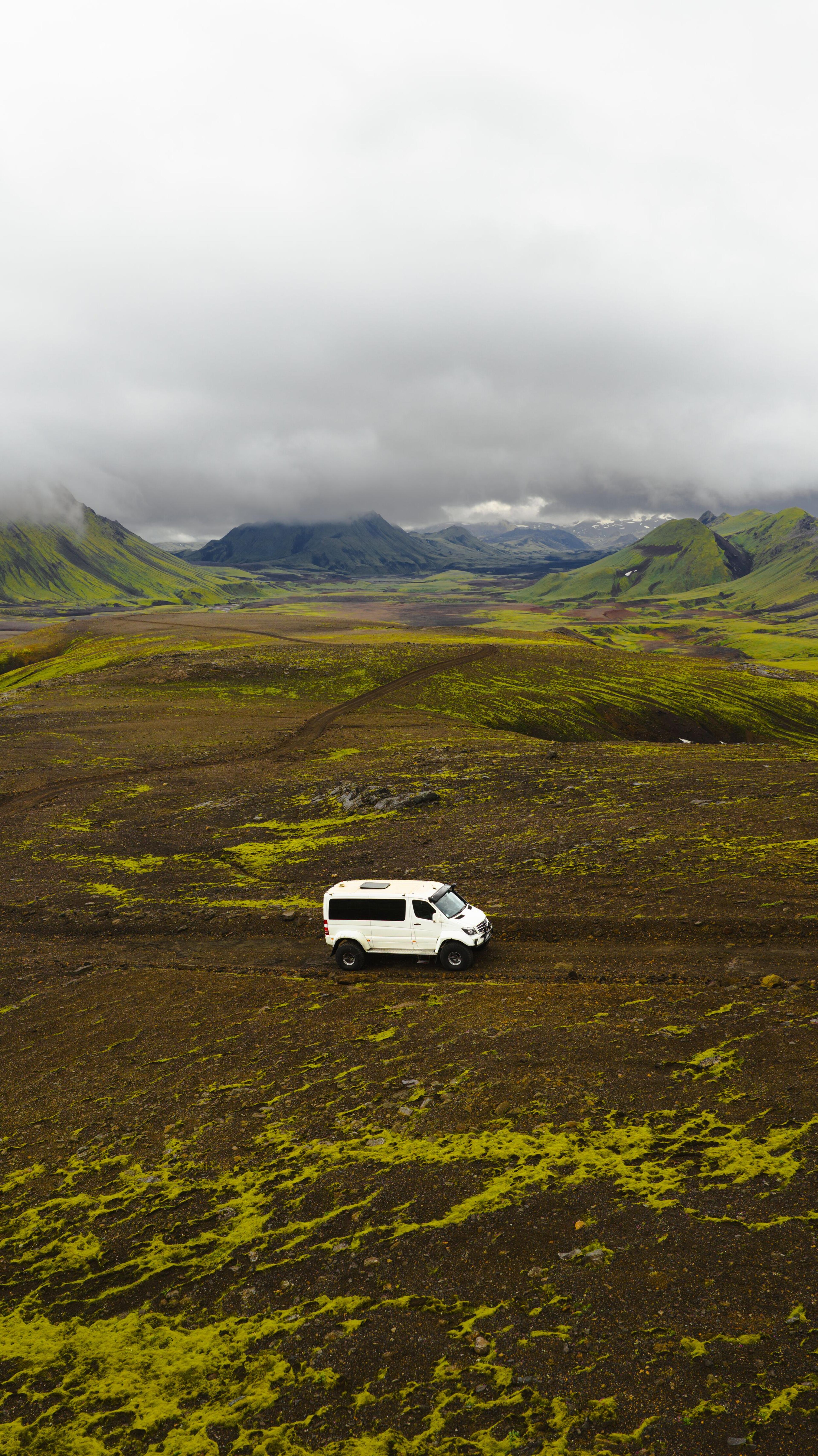 A white off-road vehicle drives on a dirt track through a vast green and brown valley with mountains under an overcast sky.