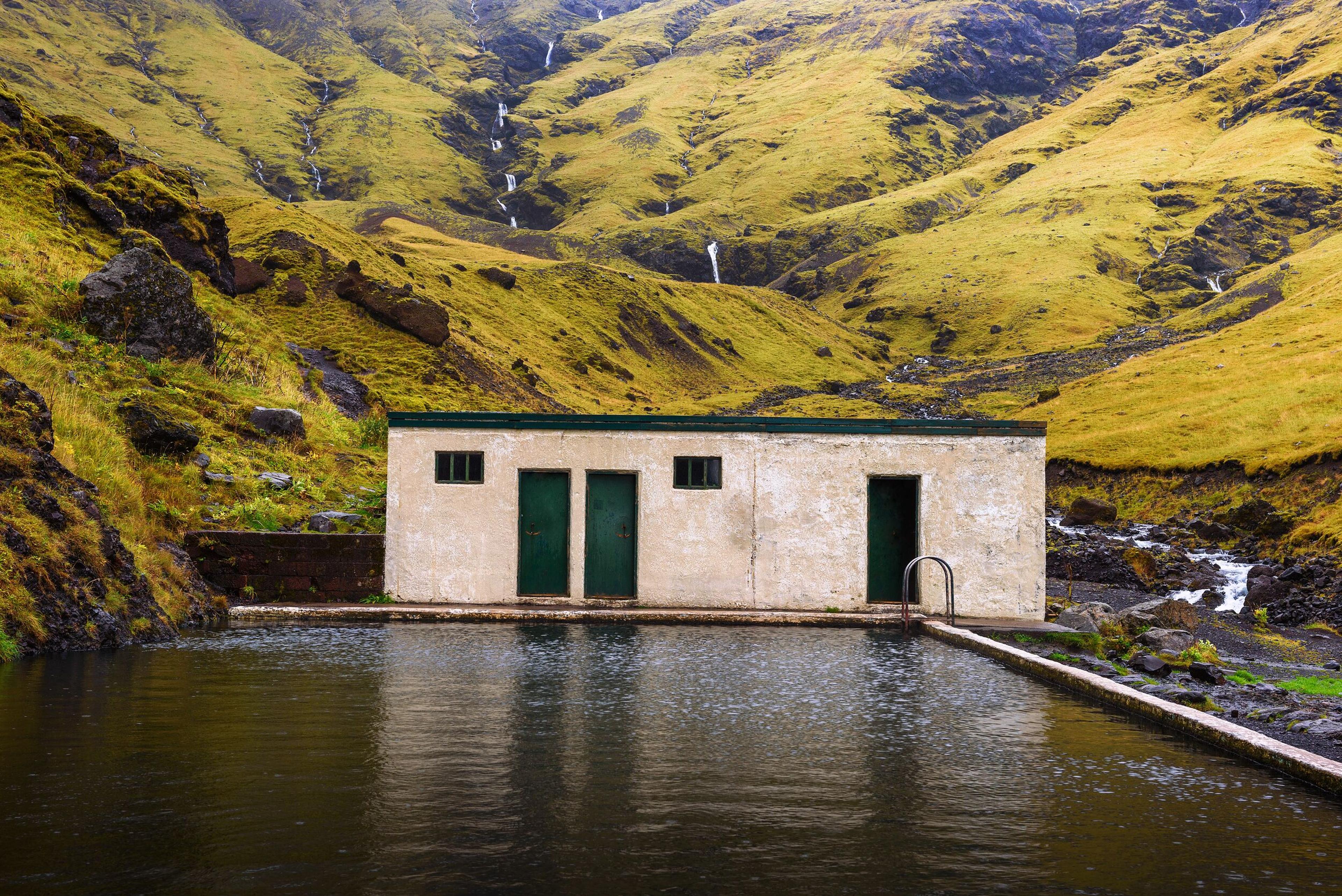 An outdoor swimming pool and a light-colored building at the base of a vibrant green, mossy hill with multiple waterfalls.