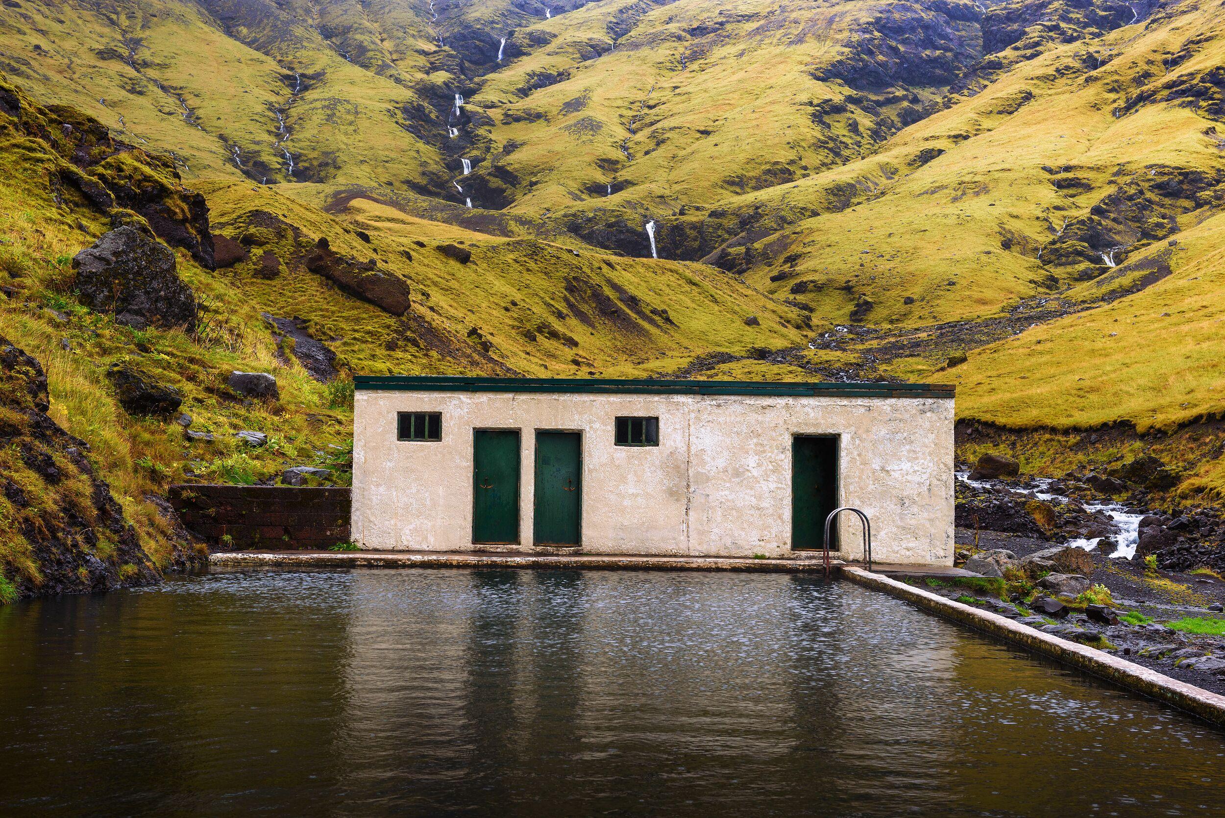 An outdoor swimming pool and a light-colored building at the base of a vibrant green, mossy hill with multiple waterfalls.