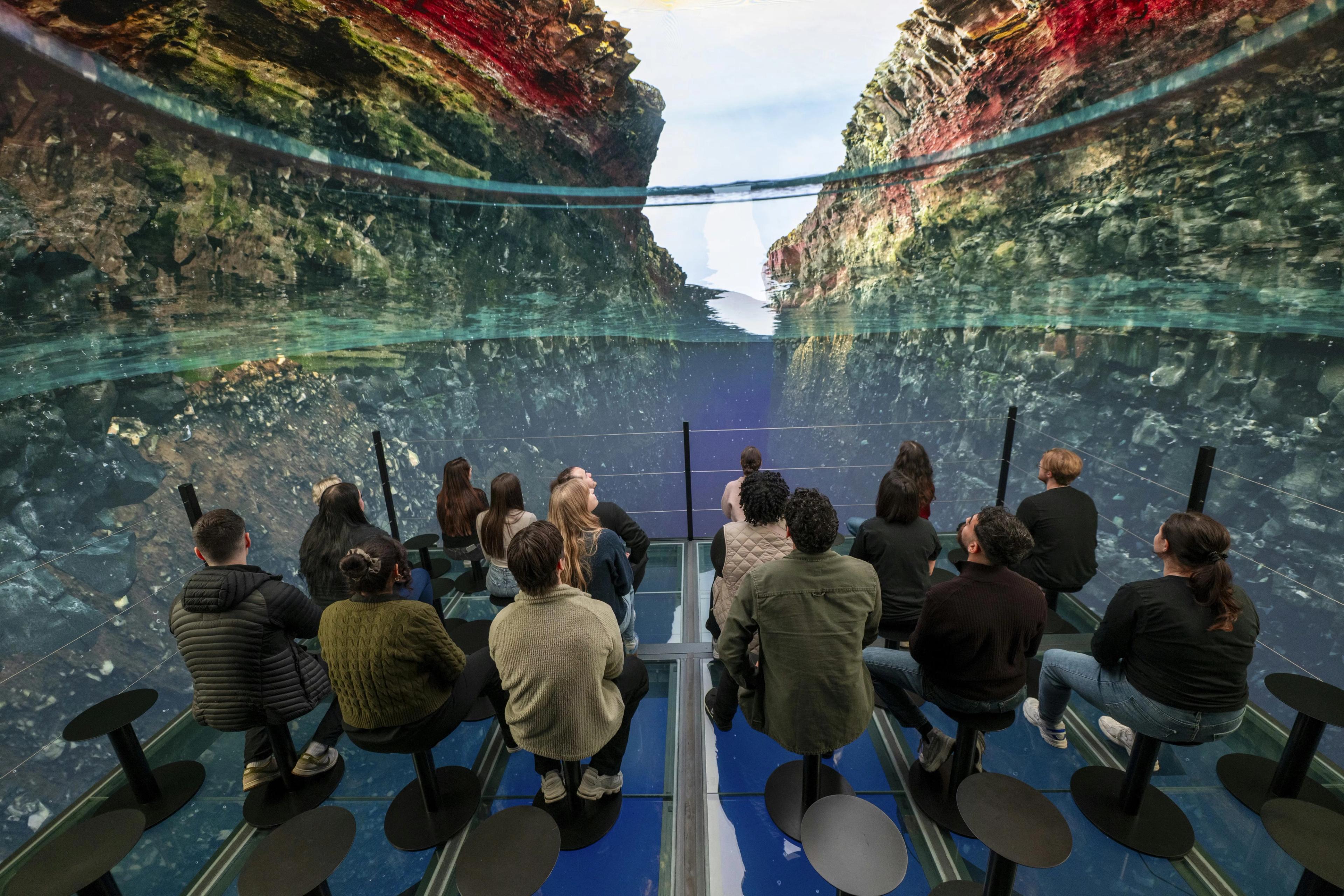 People sit on stools in an immersive exhibit, looking at a panoramic display of a deep, rocky canyon with water and reflections.