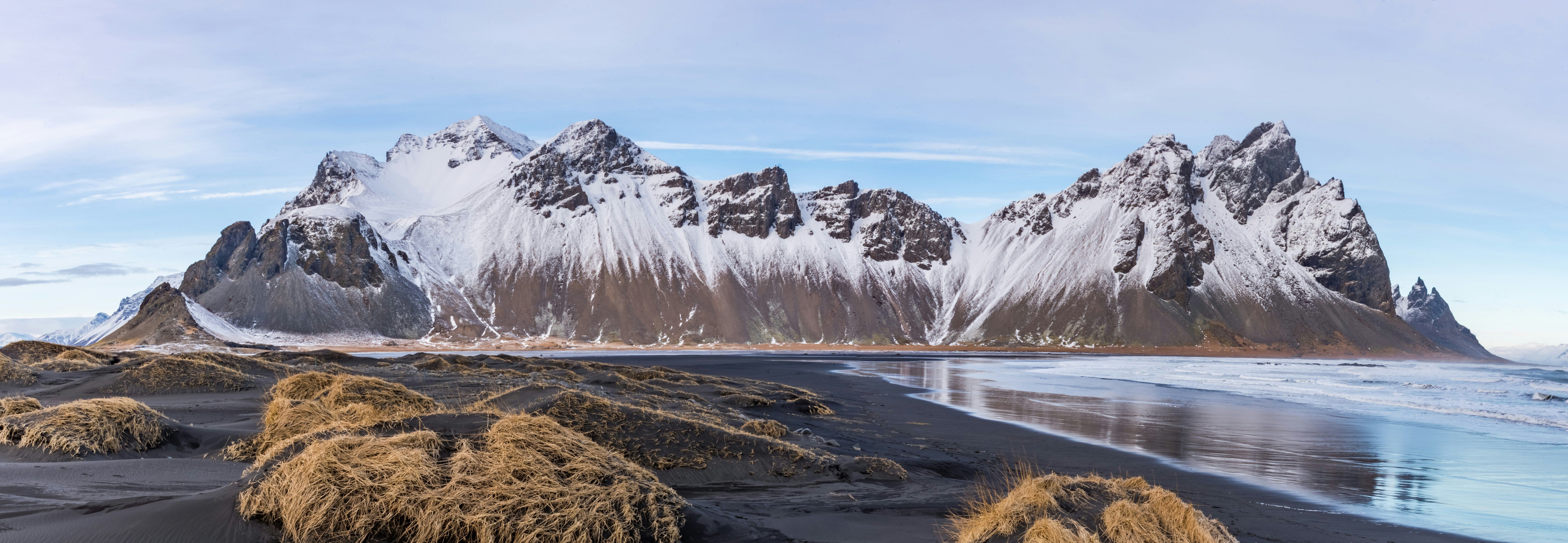 This image showcases the snow-dusted peaks of the Vestrahorn mountain reflected in the calm, wet black sands of a beach in Iceland. The serene ocean and clear blue sky add to the breathtaking contrast of dark volcanic terrain and bright snowy accents. 